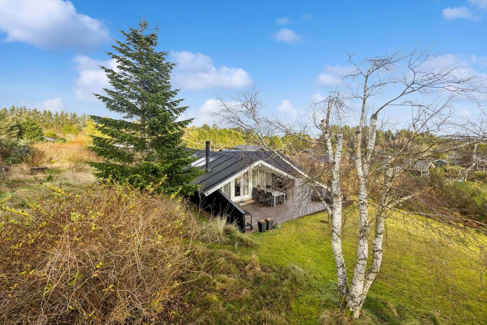 Modern wooden cabin with terrace, surrounded by trees and grass.
