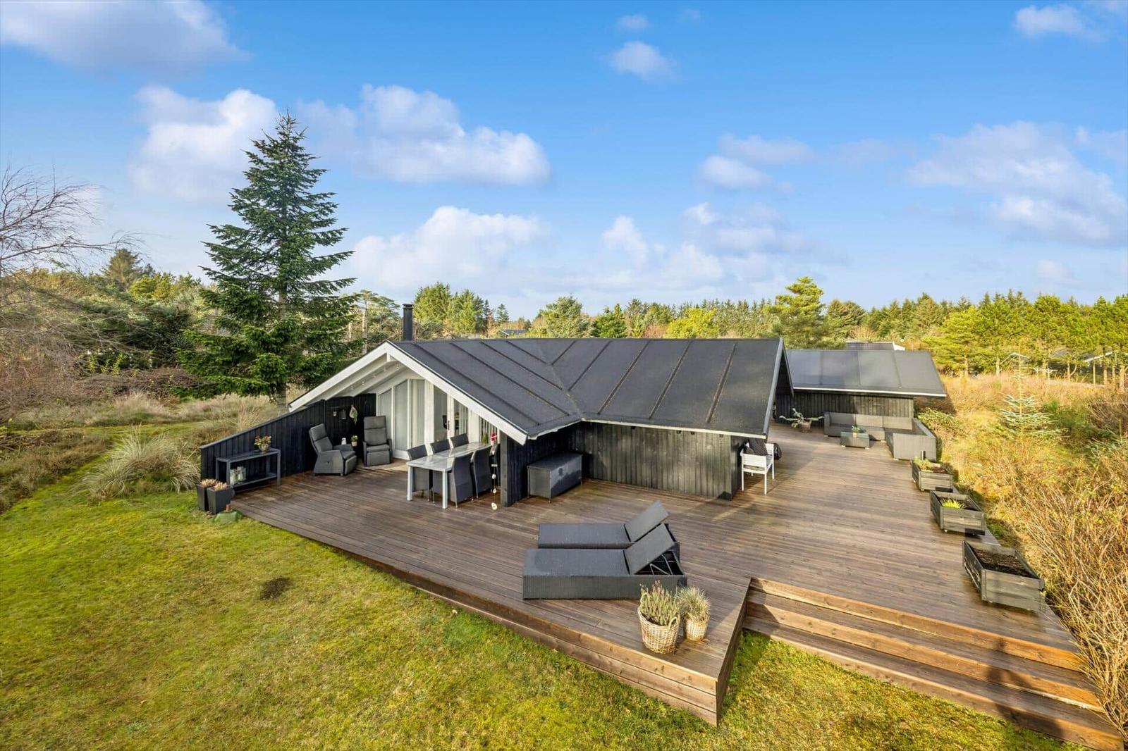 Modern villa with large wooden terrace, garden furniture, and view of forest.