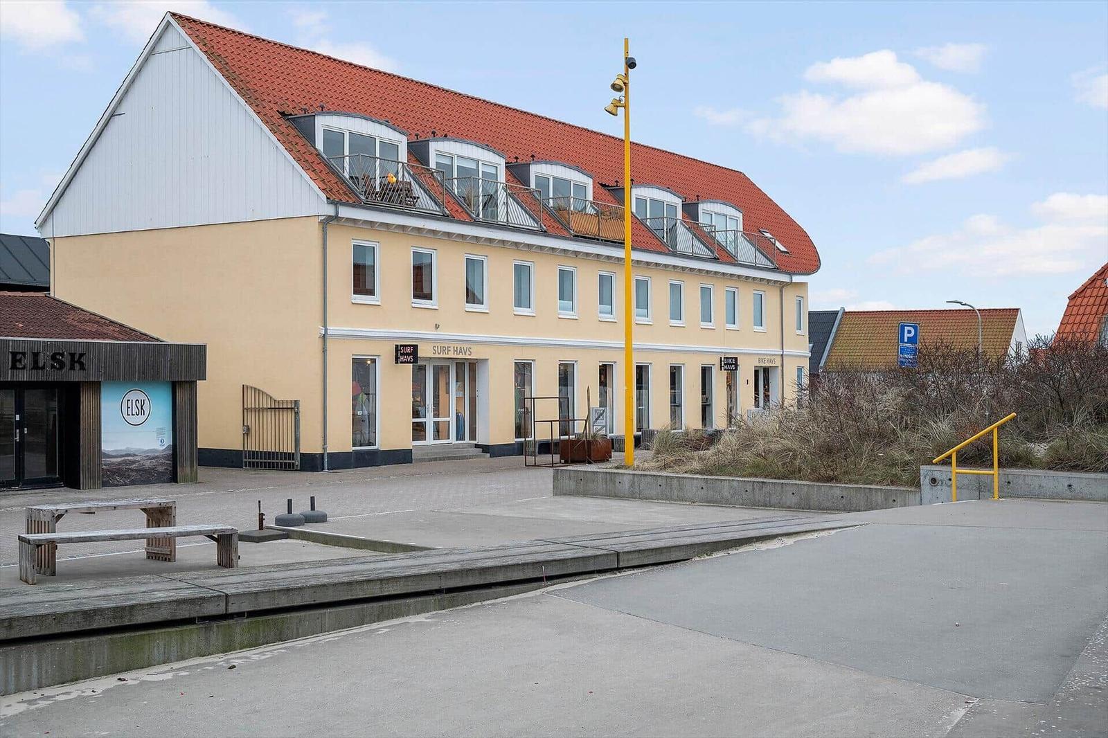 Yellow building with red roof and balconies. Front has shops and parking lot.