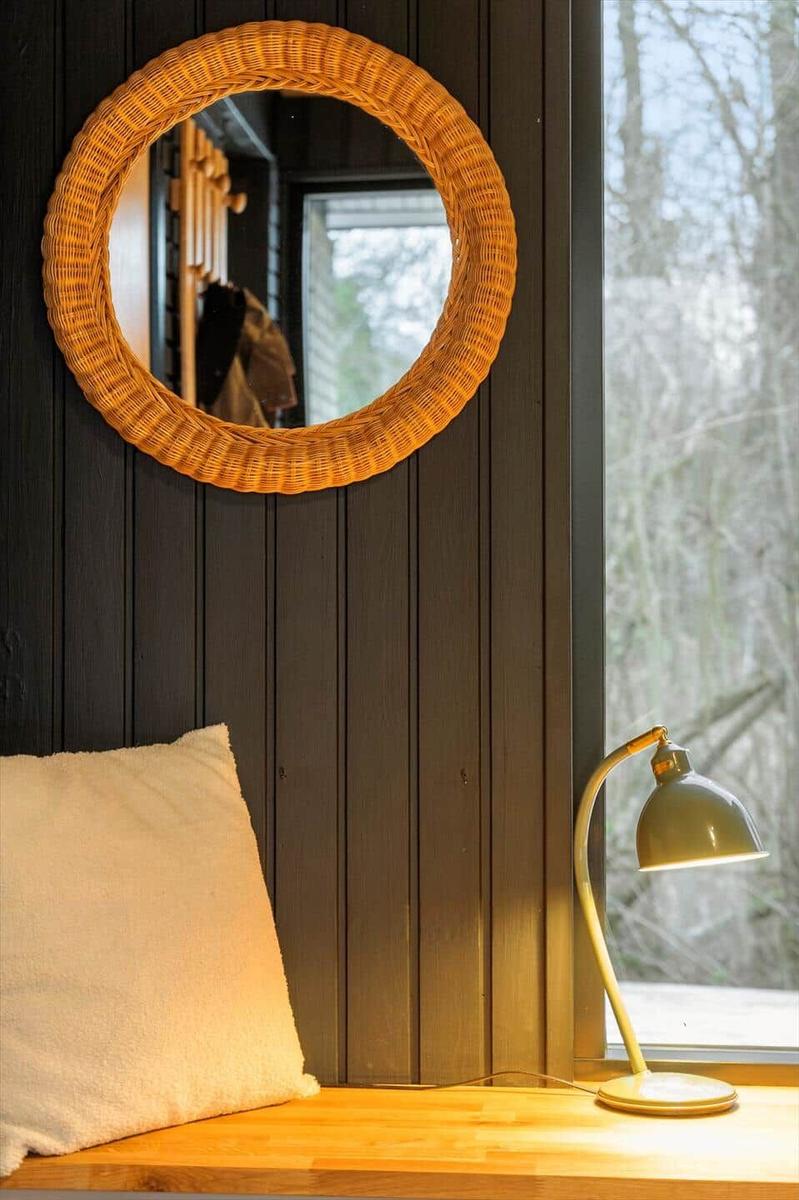 Round wicker mirror on dark wall, next to window with lamp and cushion.