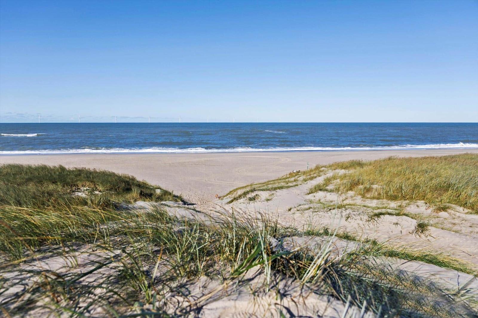 Strand mit Dünen und Windkraftanlagen im Hintergrund.