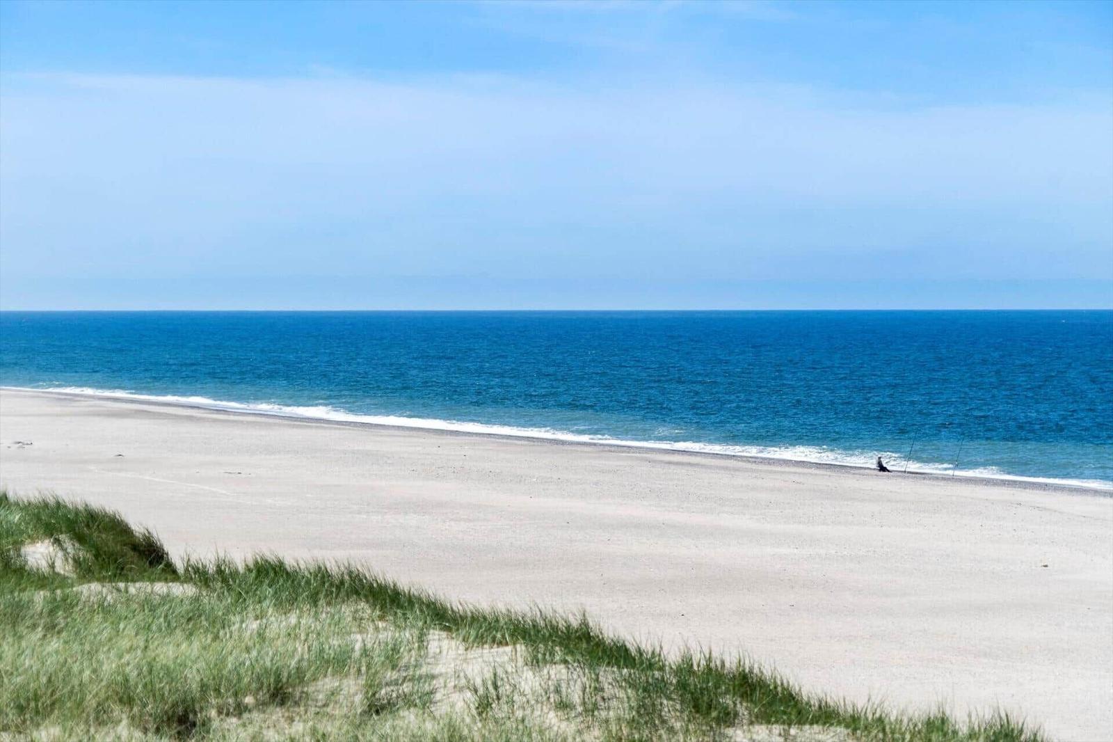 Weiterer Blick auf den Strand mit blauem Meer und Sanddünen im Vordergrund.