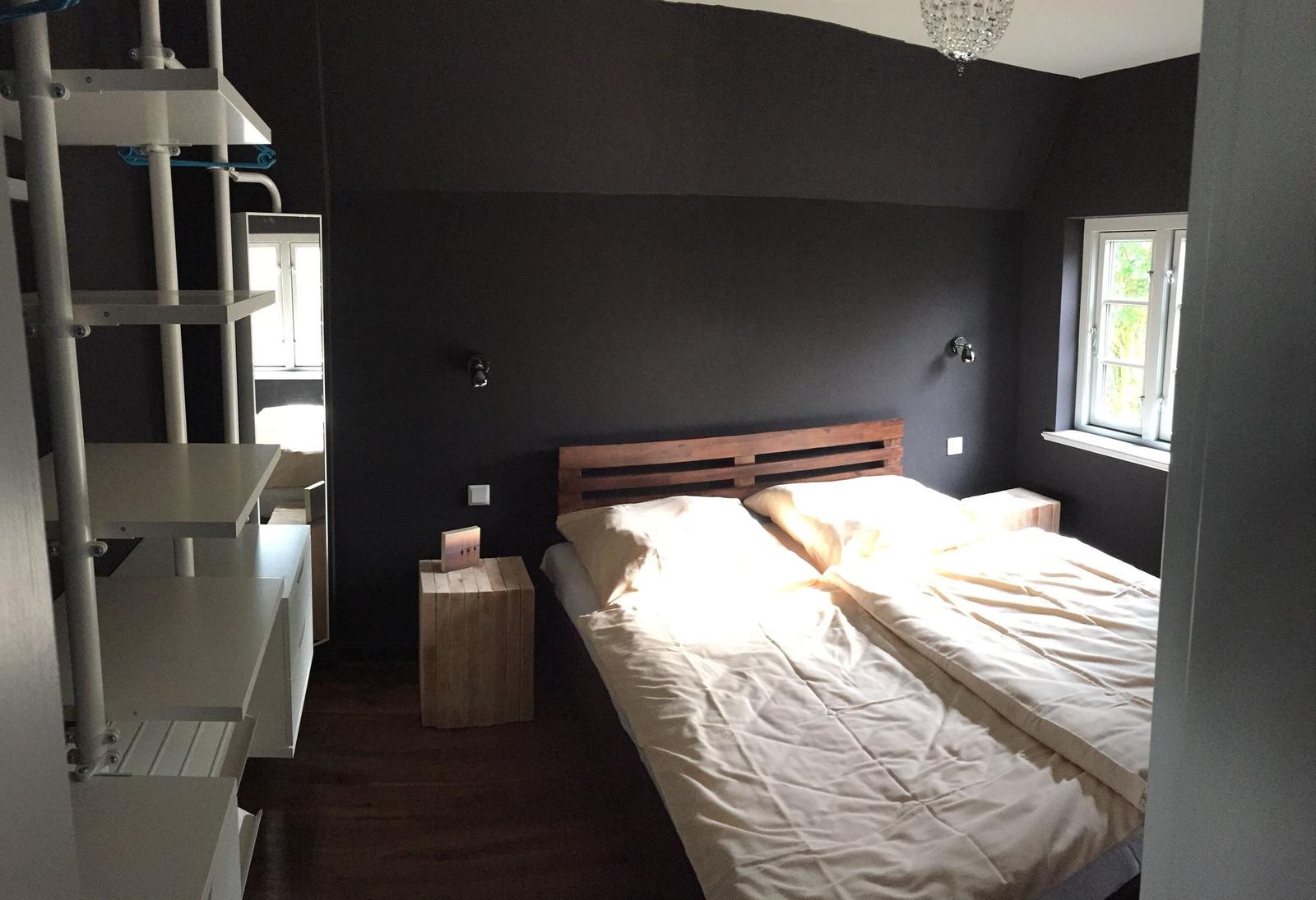 Schlafzimmer mit Bett, Nachttisch und Fenster. Dunkle Wände und Holzboden.