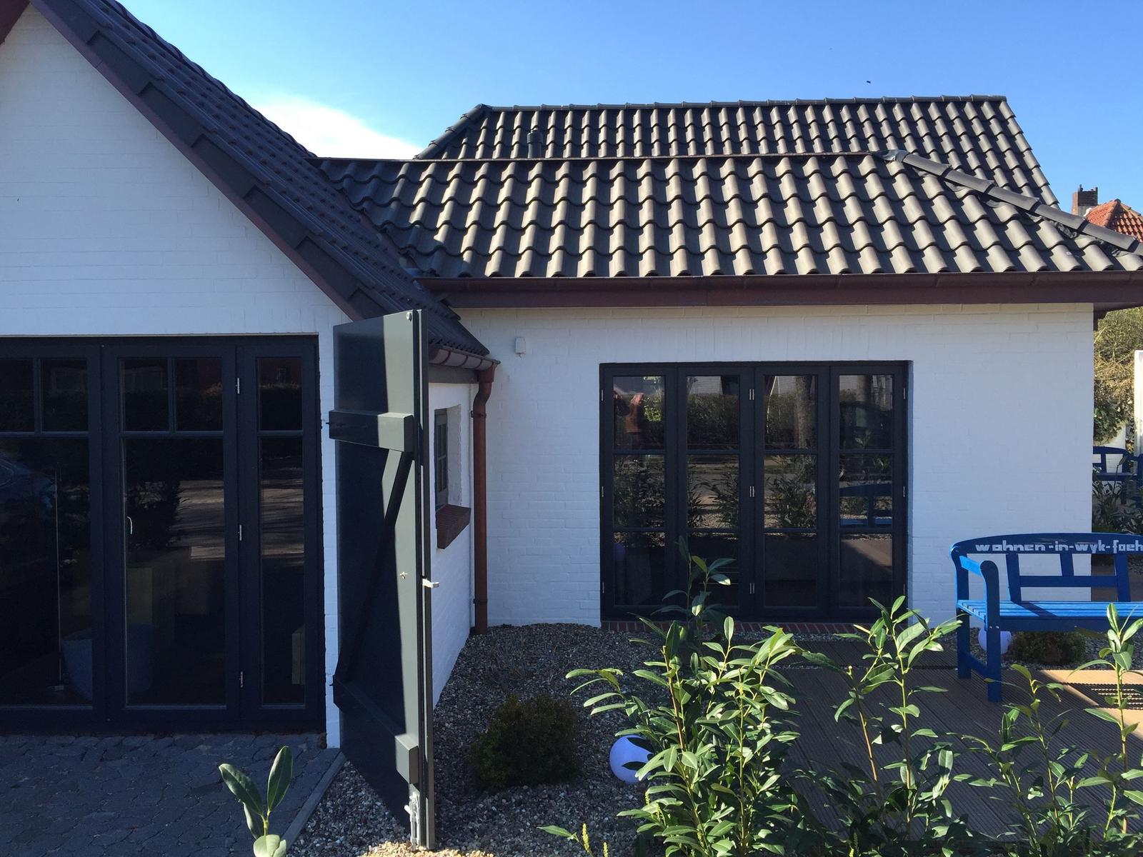 Wohnhaus mit weißen Wänden, dunklem Dach und großzügigen Fenstern. Holzterrasse mit blauer Bank.