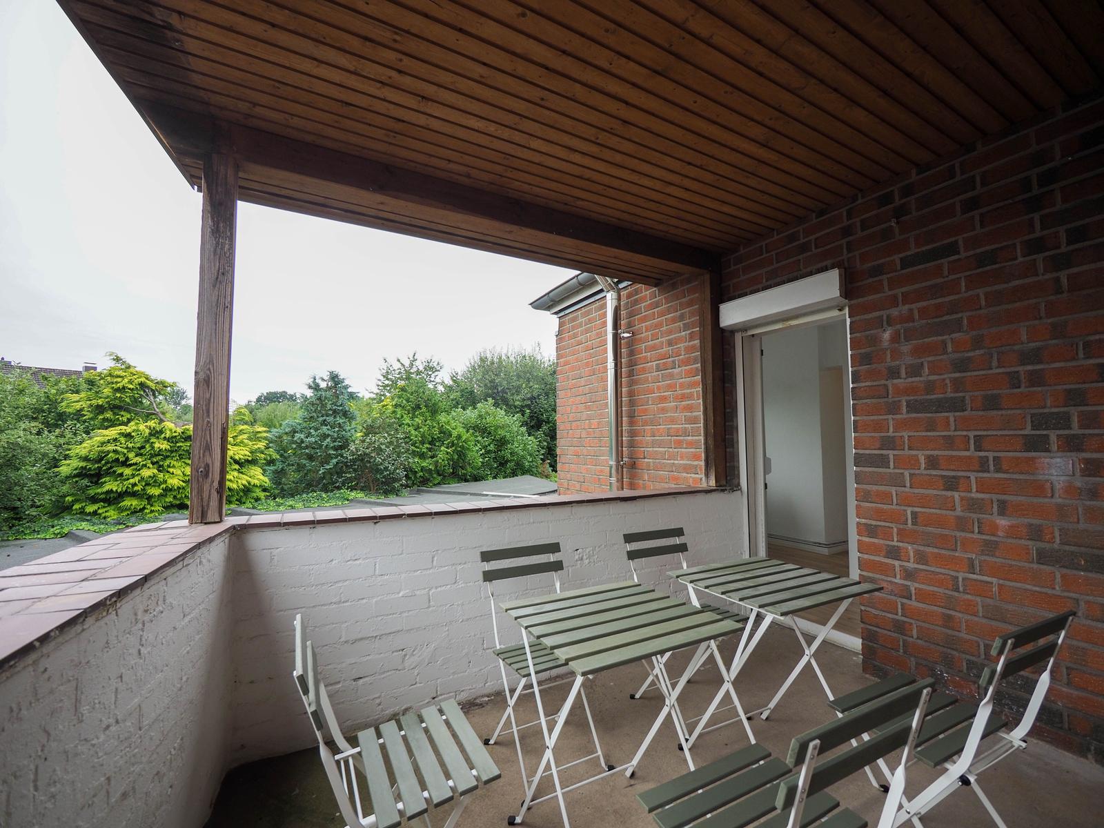 Terrasse mit Tisch und Stühlen, Blick auf grüne Bäume und roten Backsteinbau.