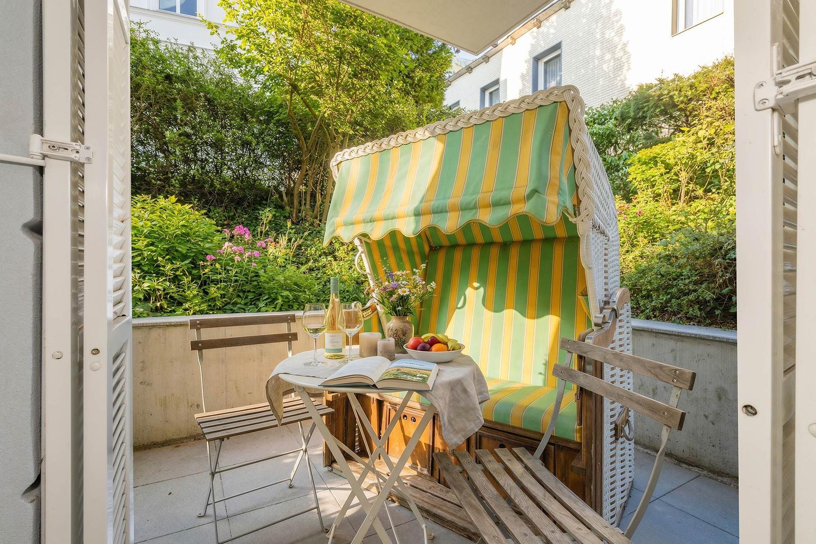 Terrasse mit Strandkorb, Tisch und Stühlen. Hintergrund: grüne Pflanzen.