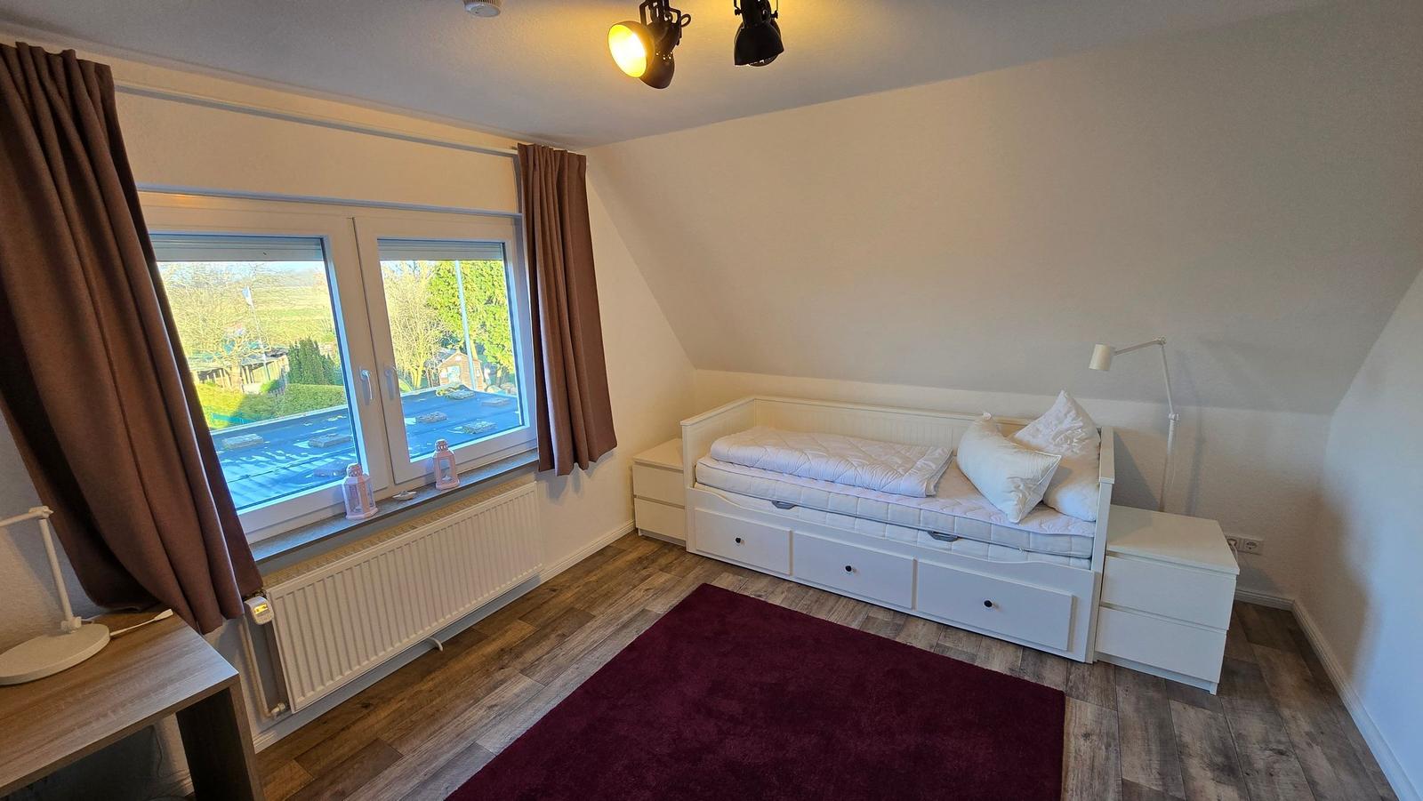 Schlafzimmer mit Dachschrägen, weißem Bett und Holzboden. Fenster mit braunen Vorhängen.