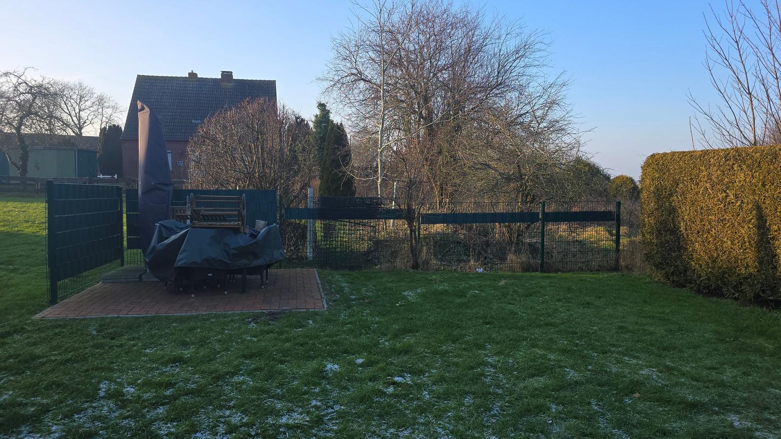 Hinterhof mit Rasenfläche, Terrasse und Gartenzaun. Gartenmöbel sind zugedeckt.