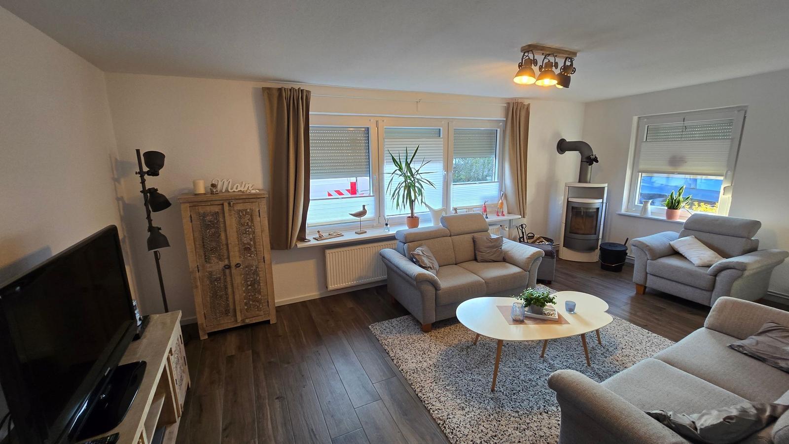 Wohnzimmer mit Sofa, Sessel, Couchtisch und Kaminofen. Holzboden und Fenster mit Jalousien.