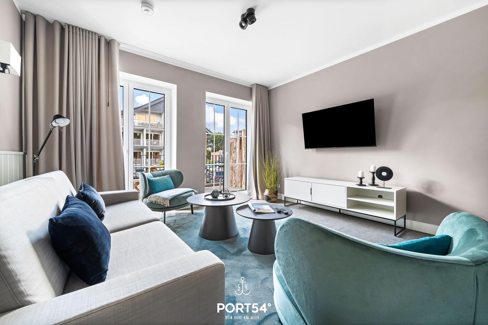 Wohnzimmer mit Sofa, Sesseln, TV und Fenstern zum Balkon.