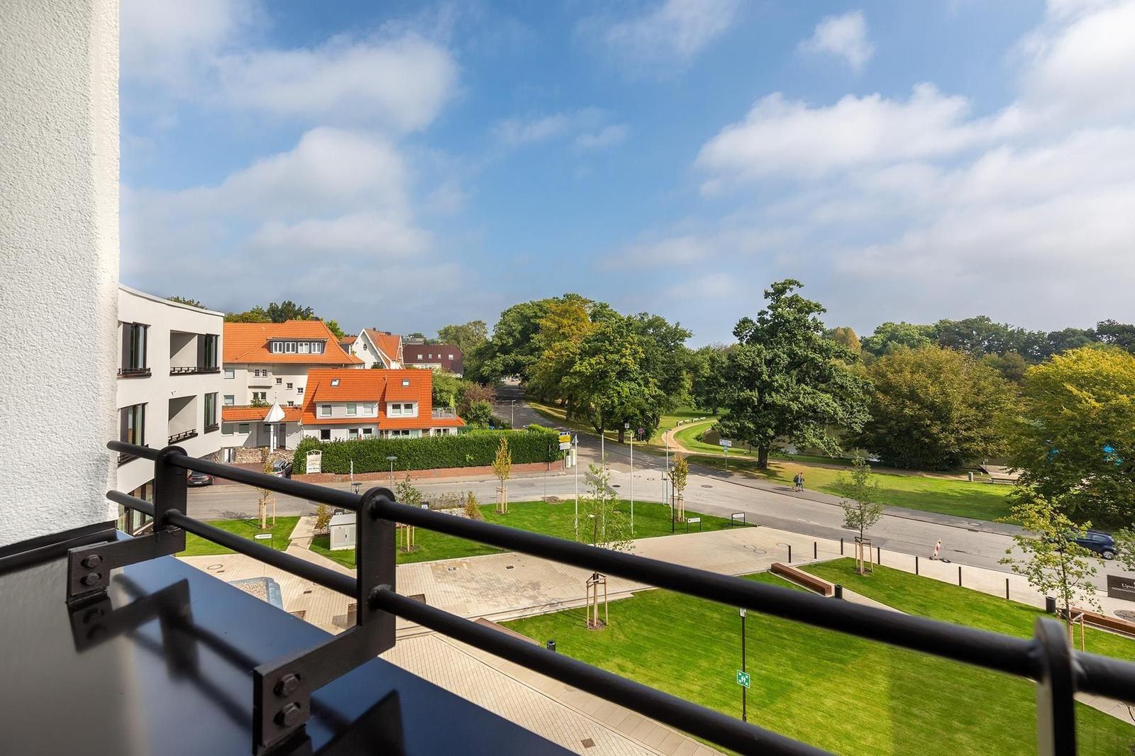 Blick vom Balkon auf Grünflächen, Straßen und Gebäude unter blauem Himmel.