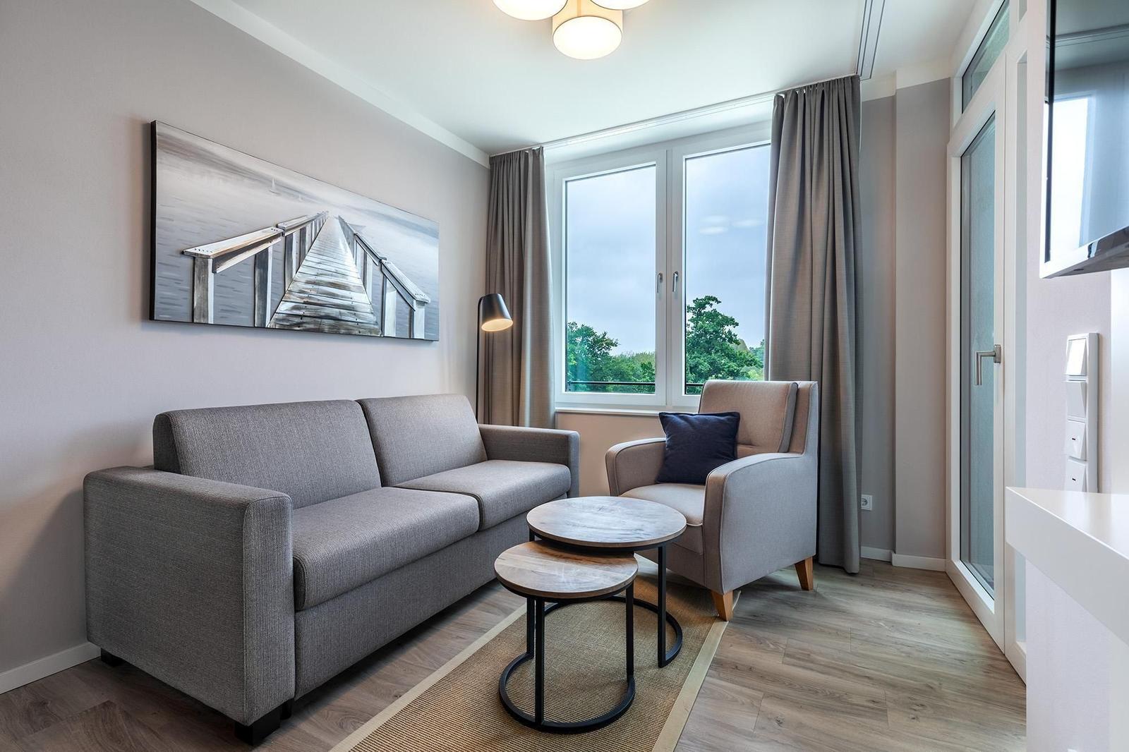 Wohnzimmer mit Sofa, Sessel und Tisch. Fenster mit Blick auf Grün. Wandbild und Deckenleuchte.