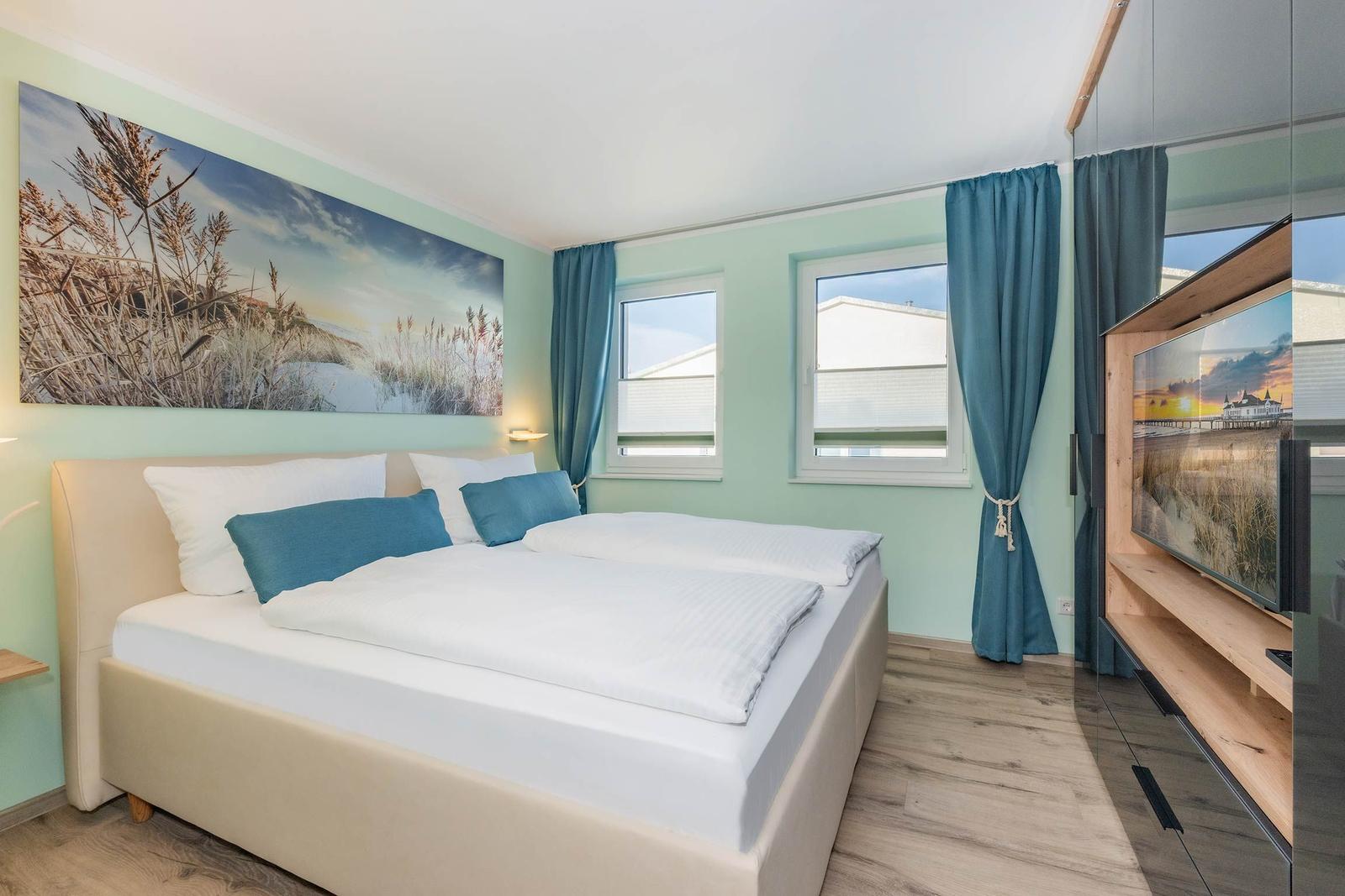 Schlafzimmer mit Doppelbett, Fenstern, Vorhängen und Fernseher.