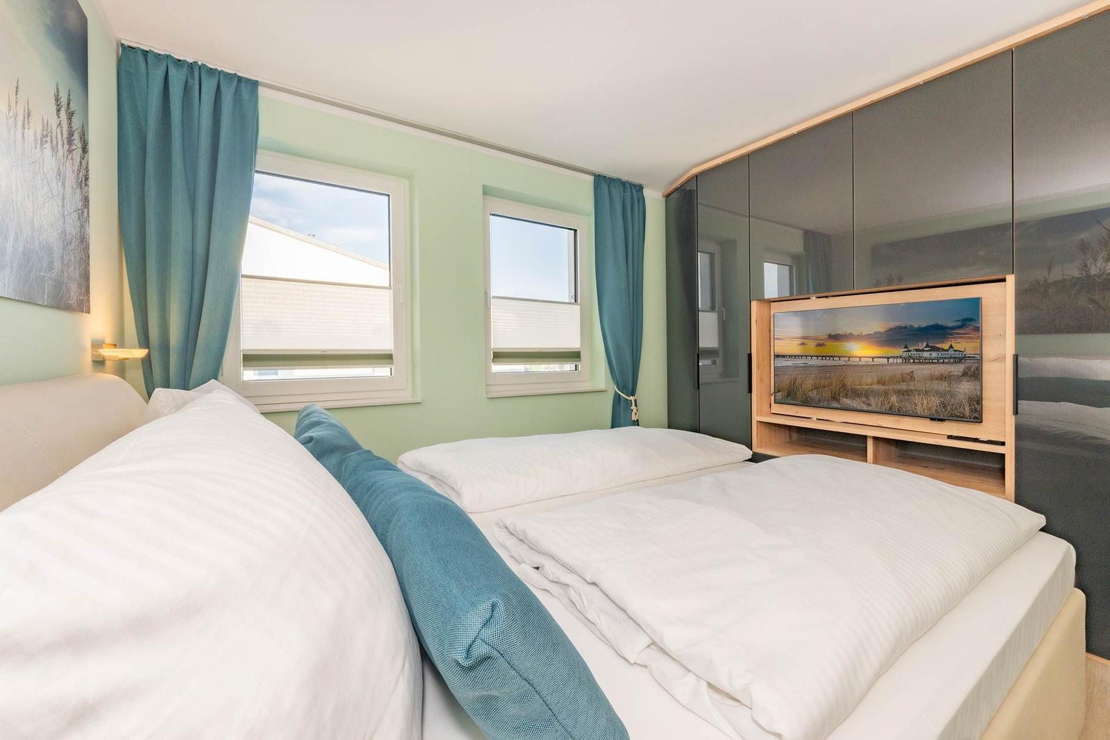 Schlafzimmer mit Doppelbett, zwei Fenstern und TV im Schrank.