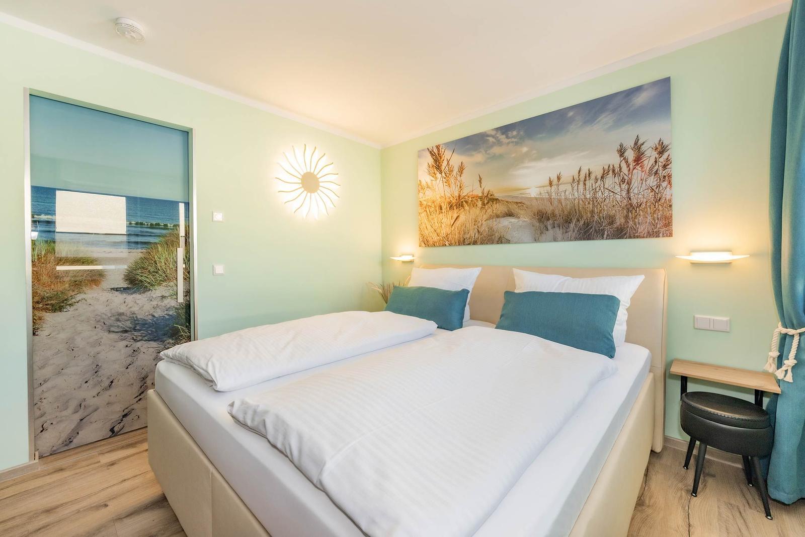 Schlafzimmer mit Bett, Wandbild und Tür zur Strandlandschaft.