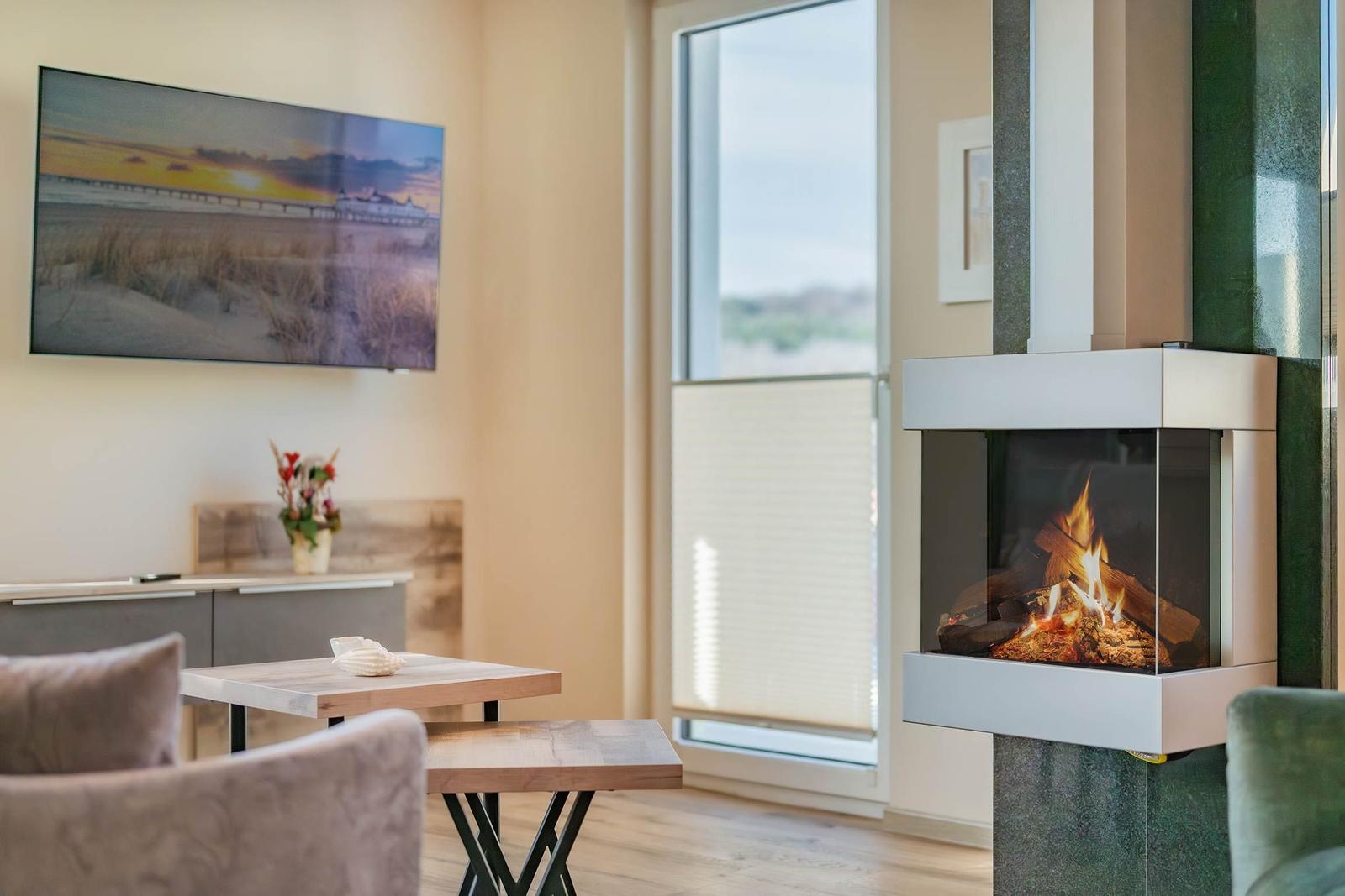 Wohnzimmer mit Kamin, TV und Tisch. Große Fenster mit Jalousien.