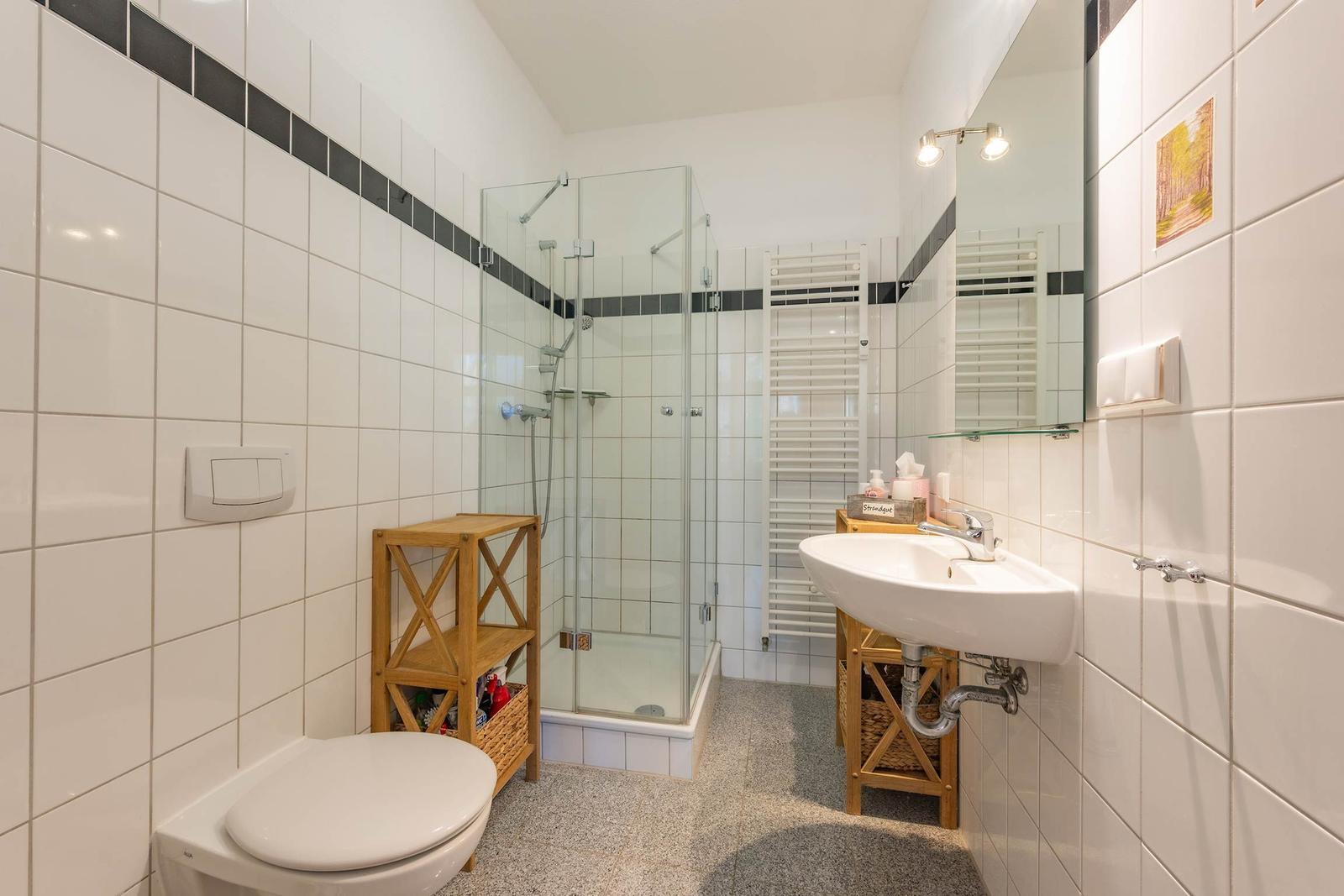 Badezimmer mit Dusche, Waschbecken und Toilette. Weiße Fliesen und Holzregale.