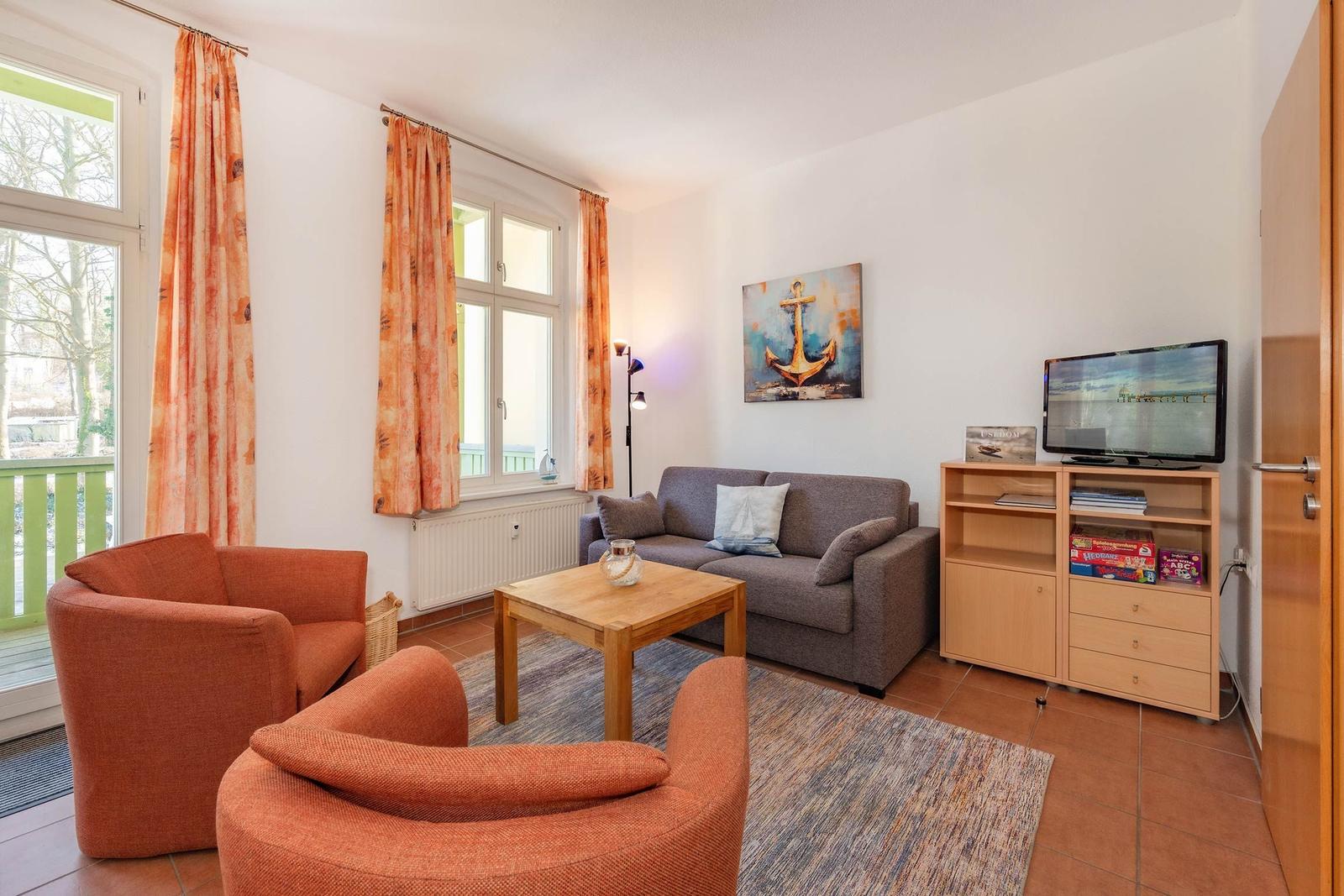 Wohnzimmer mit Sofa, zwei Sesseln, TV und Fenstern mit Vorhängen.