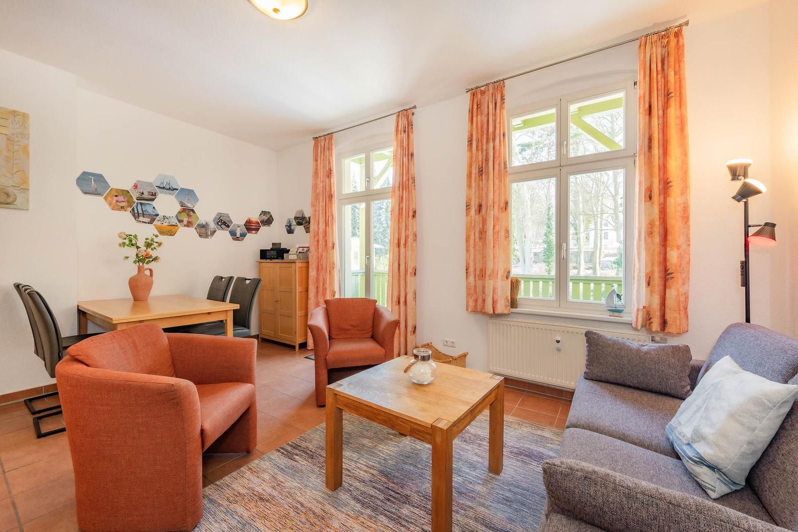 Wohnzimmer mit Sofa, Sesseln, Tisch und Fenstern mit Vorhängen.