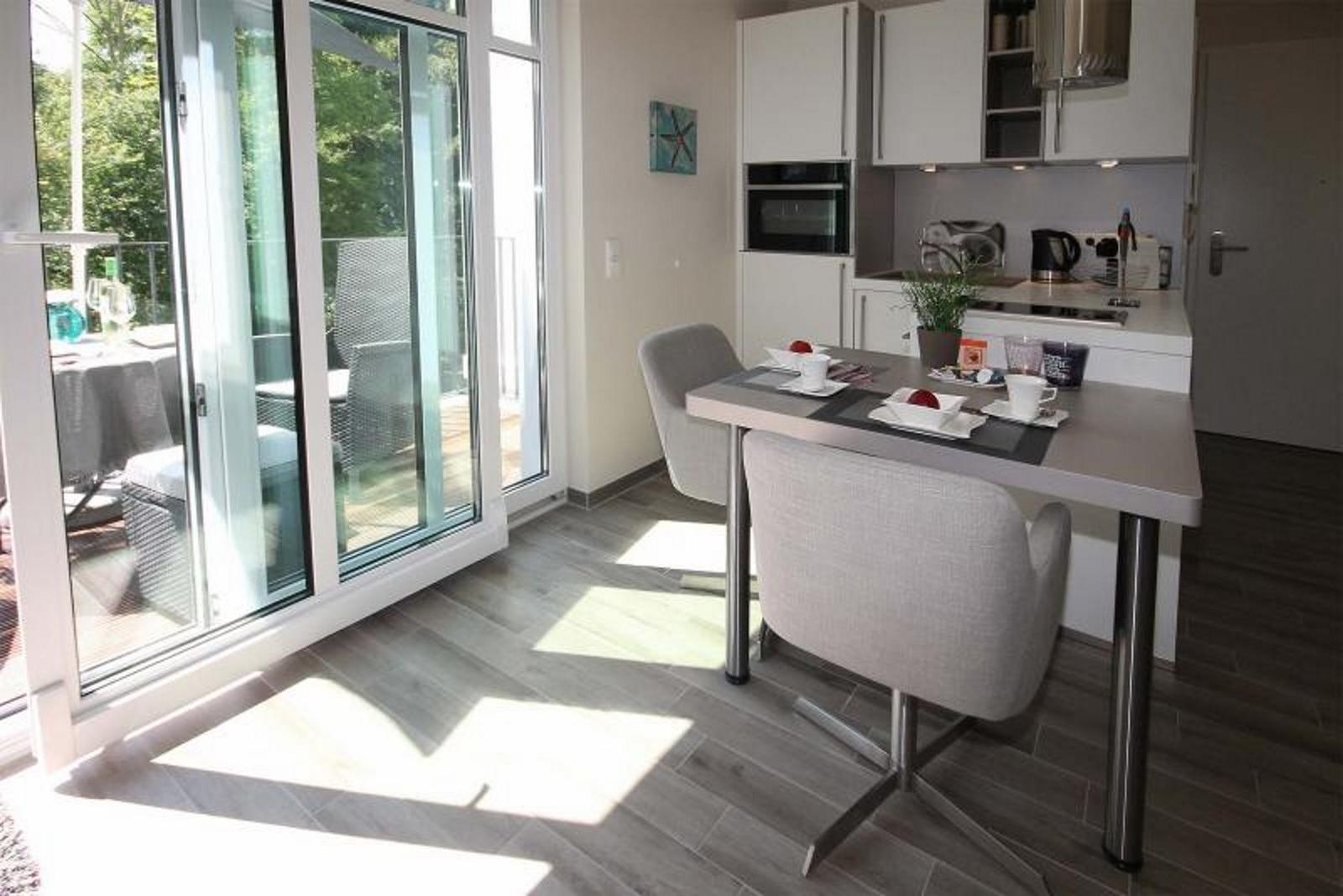 Essbereich mit Tisch, Stühlen und Blick auf Balkon. Küche mit weißen Schränken und Arbeitsplatte.