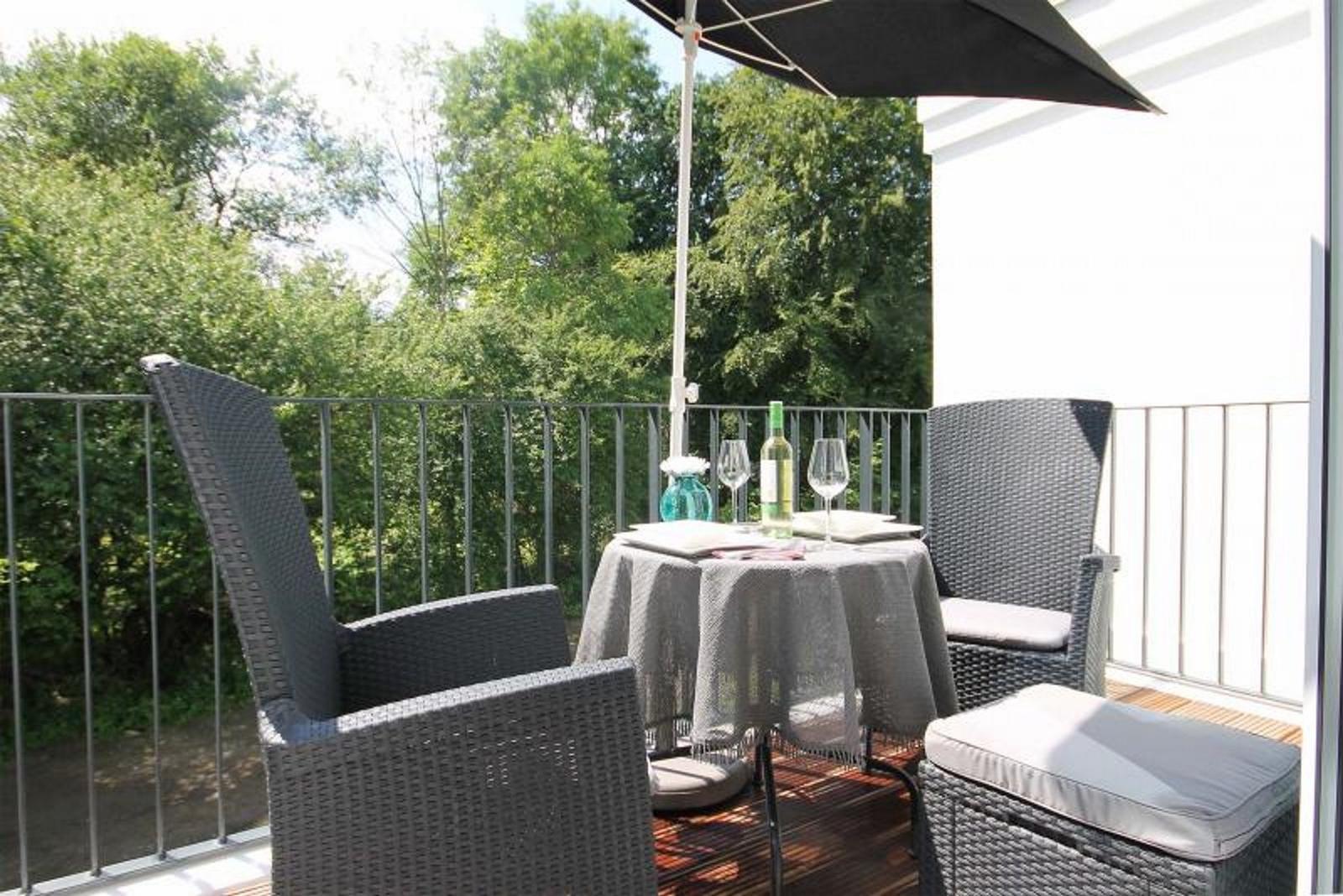 Terrasse mit Tisch, zwei Stühlen und Sonnenschutz. Hintergrund: grüne Bäume.