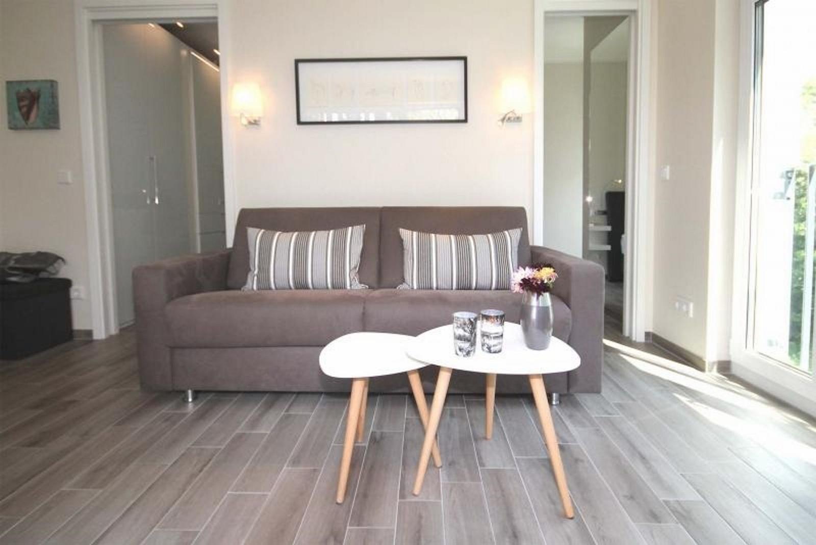 Ein Wohnzimmer mit grauem Sofa, zwei weißen Tischen und Holzboden.