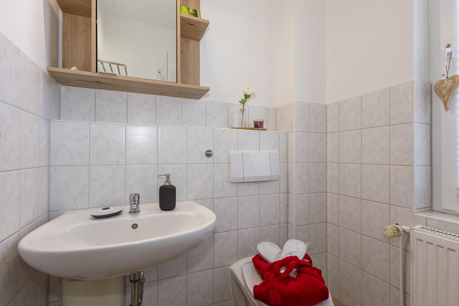 Bad mit Waschbecken, Spiegel und Toilette. Weiße Fliesen und Holzregal.