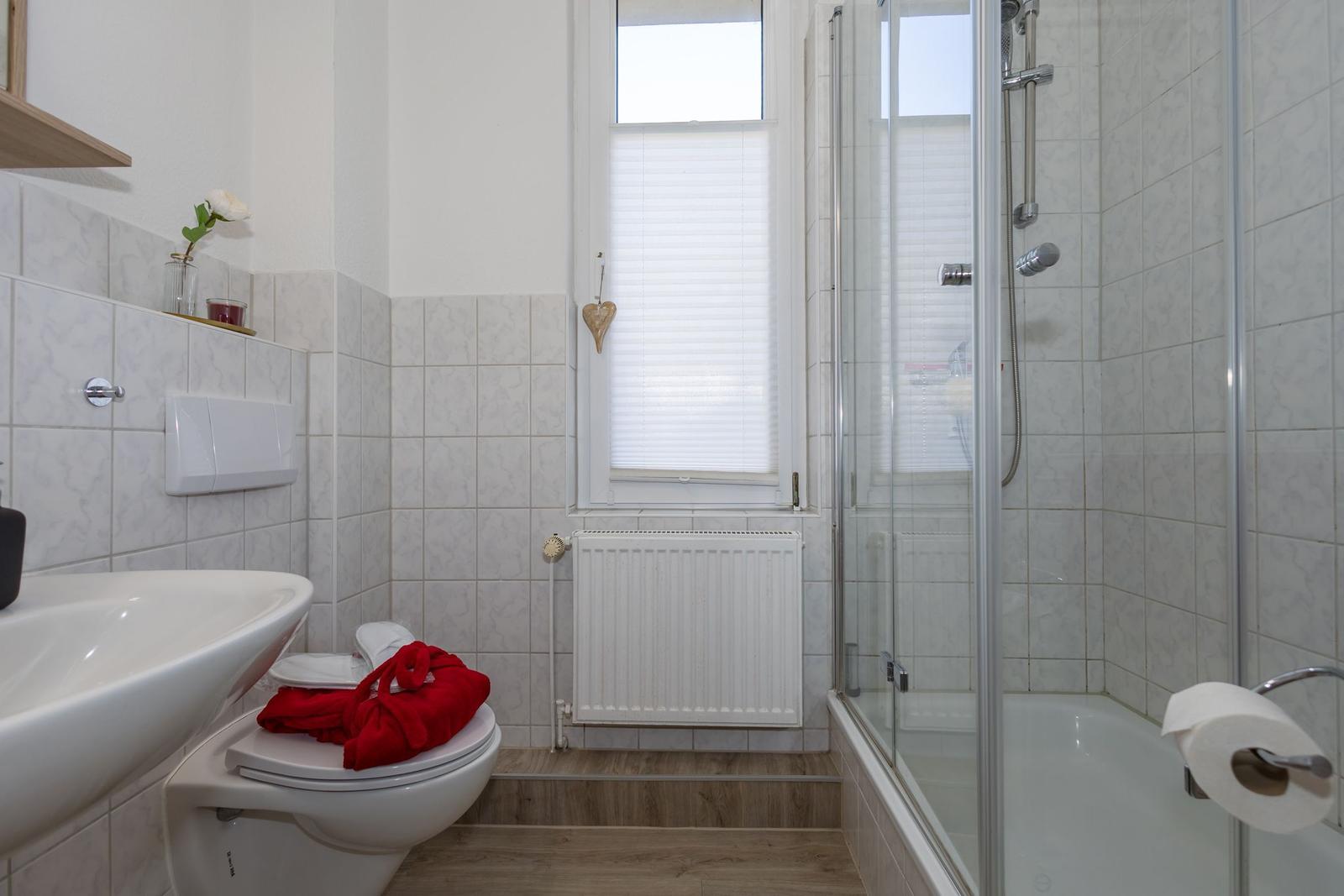 Badezimmer mit Dusche, Toilette und Waschbecken. Fenster mit Jalousie und Heizkörper.