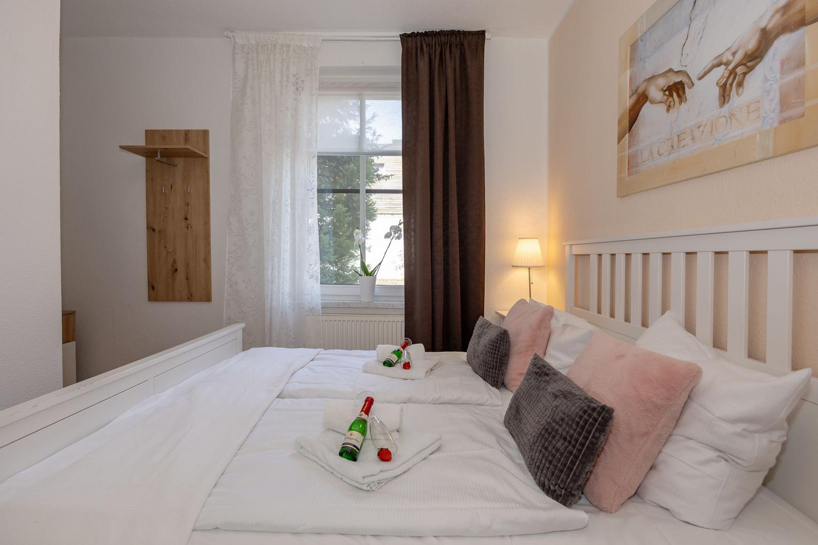 Schlafzimmer mit Bett, Fenster, Wandbild und Flaschen auf Bett.