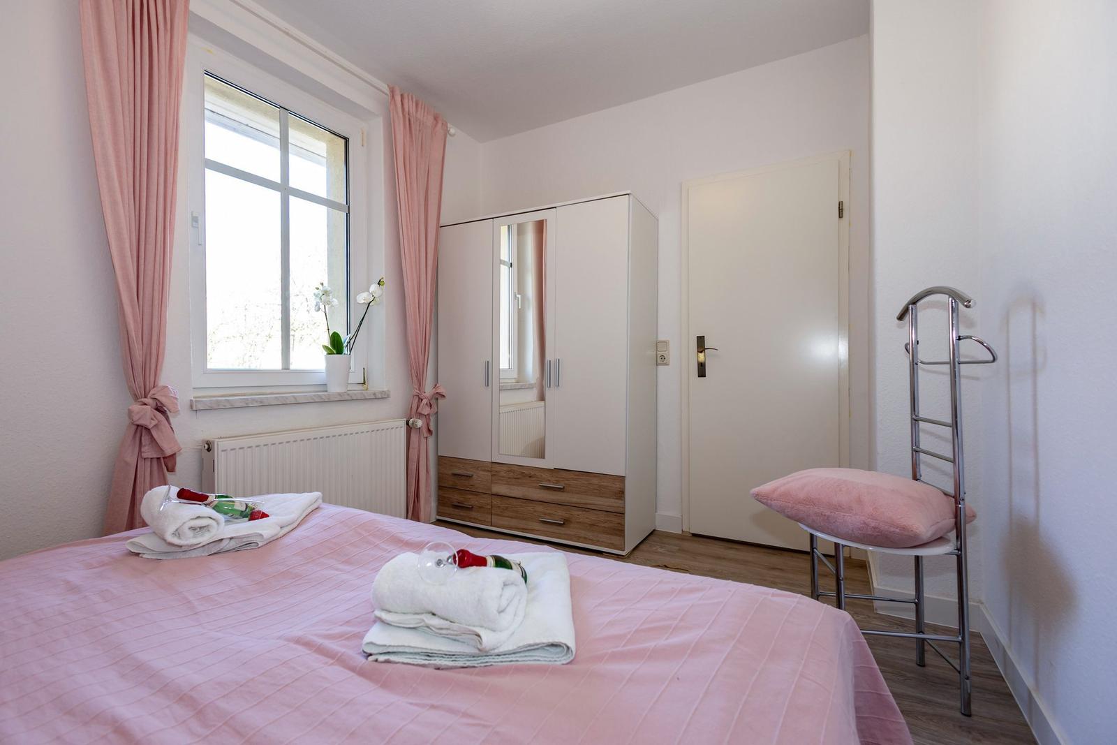 Schlafzimmer mit rosa Bettwäsche, Fenster, Kleiderschrank und Stuhl.