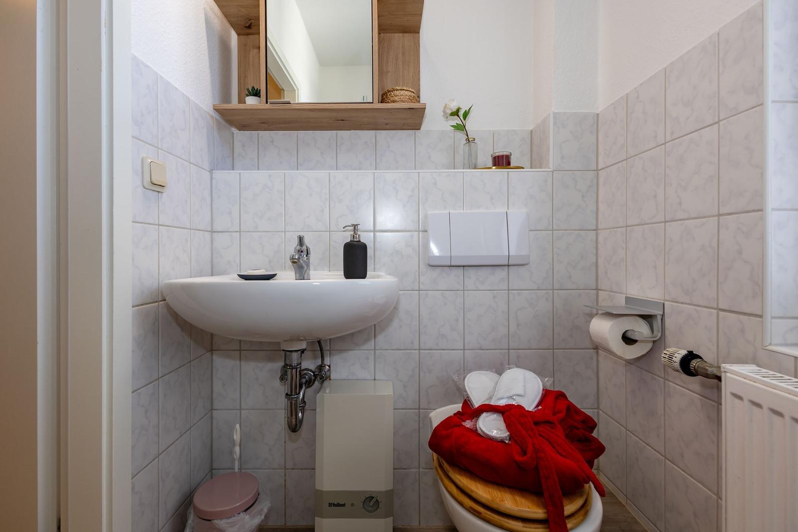 Badezimmer mit Waschbecken, Spiegel und Toilette. Auf dem WC liegt ein roter Bademantel.