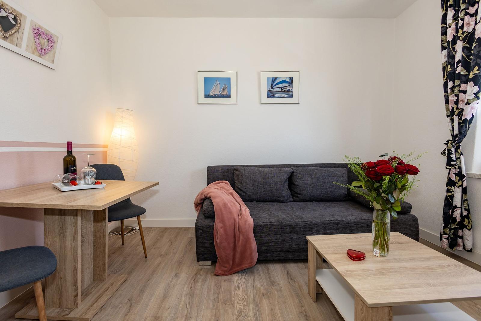 Ein Wohnzimmer mit Sofa, Tisch und Blumen. Wandbilder und Vorhänge sind sichtbar.