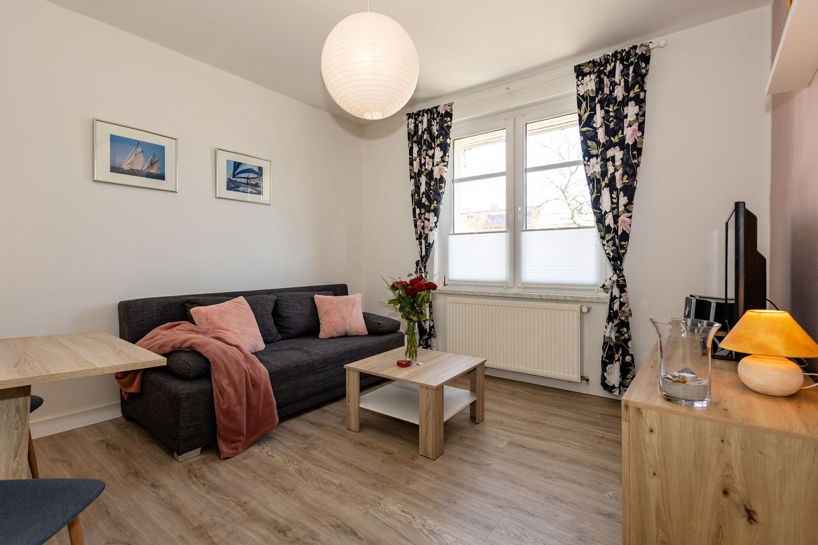Wohnzimmer mit Sofa, Tisch und Fenster mit floralen Vorhängen.