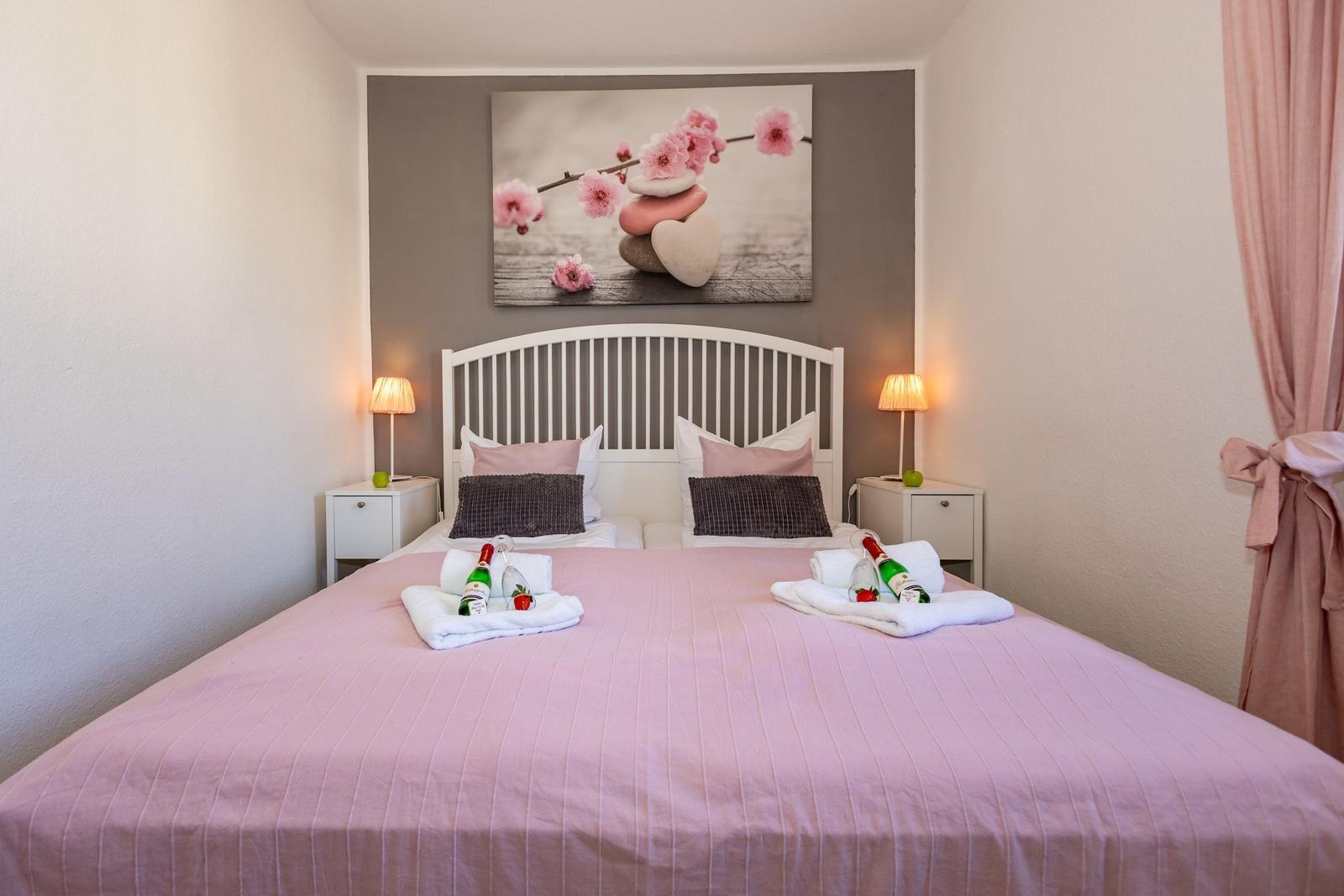 Schlafzimmer mit Bett, Nachttischen und Wandbild. Auf Bett liegen Getränke und Handtücher.