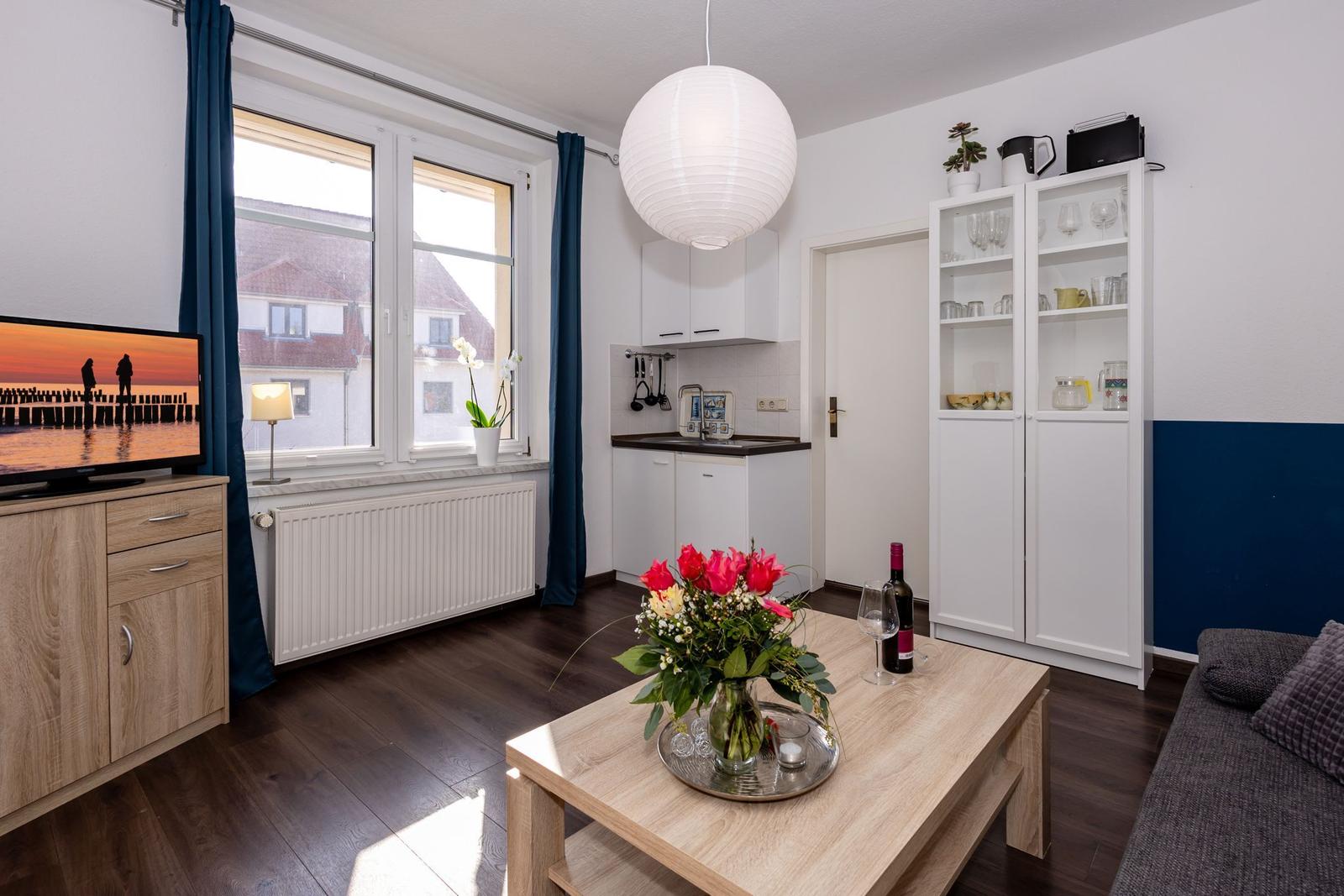 Wohnbereich mit Küche, Sofa, Tisch und TV. Fenster mit Blick auf Gebäude.