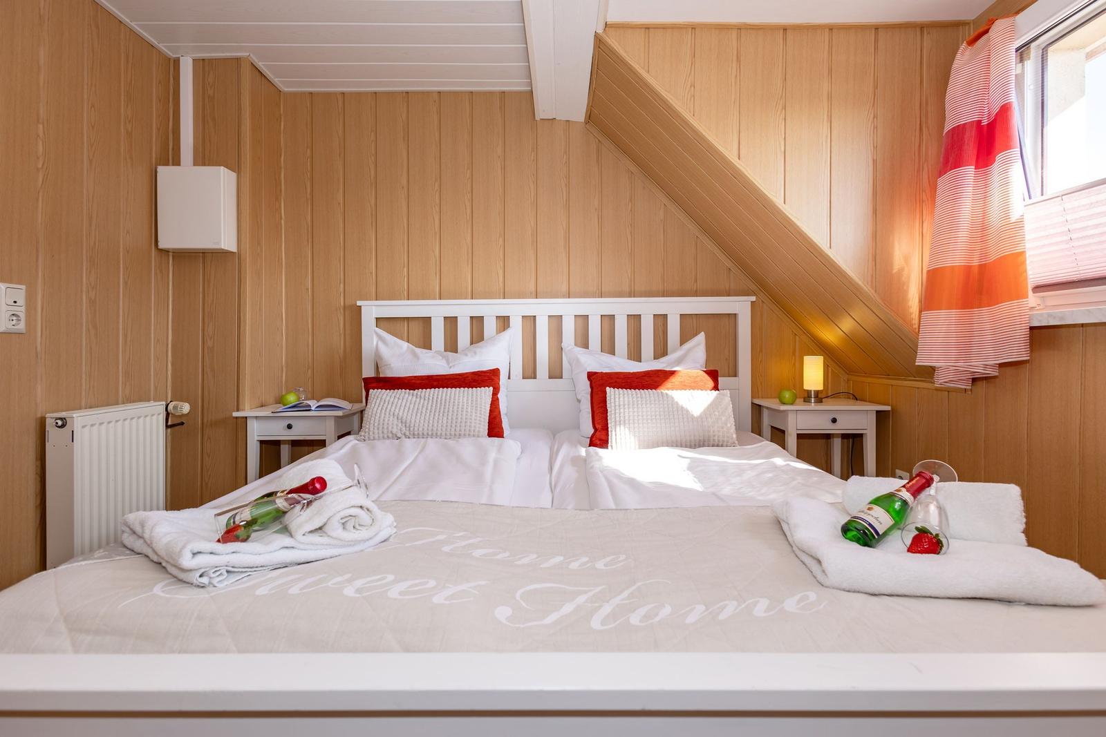Schlafzimmer mit Doppelbett, Holzwand und Fenster. Auf Bett liegen Handtücher und eine Flasche.
