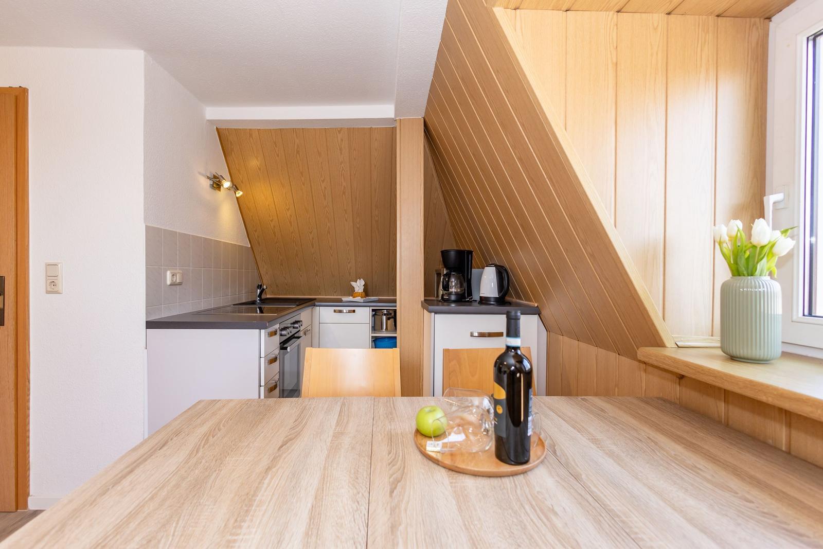 Küche mit Essbereich unter Holzdecke. Tisch mit Wein und Apfel. Fenster mit Tulpen.