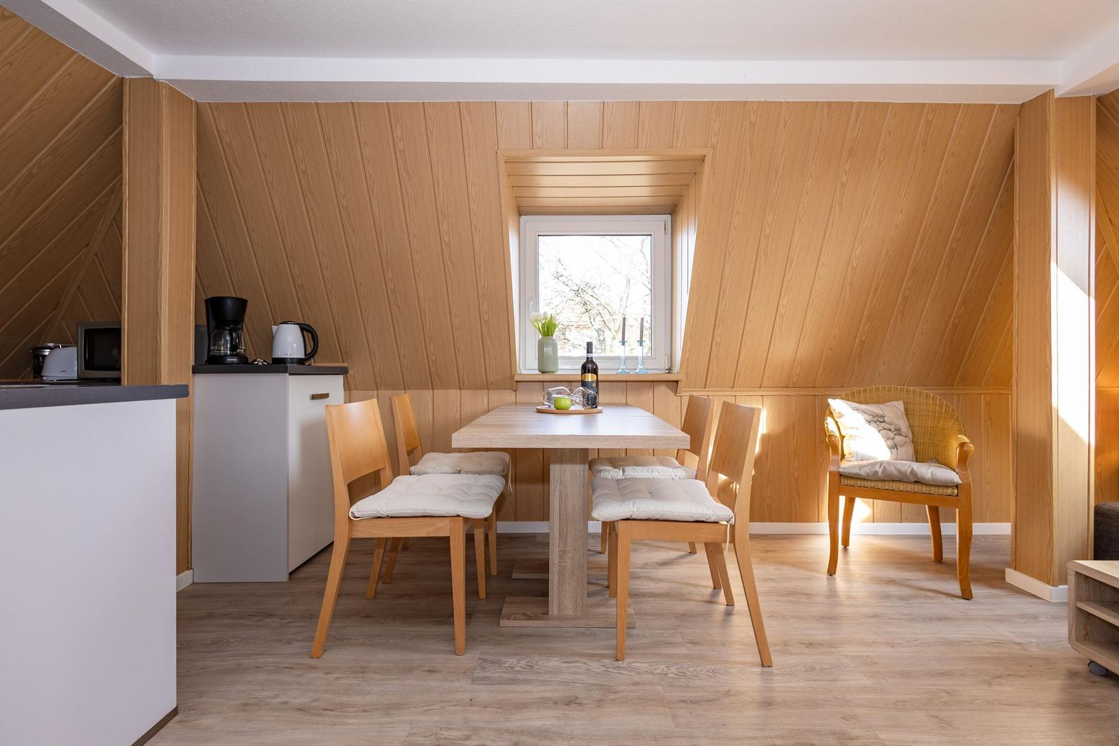 Essbereich mit Holztisch, Stühlen und Fenster. Küche mit Kaffeemaschine und Kühlschrank.