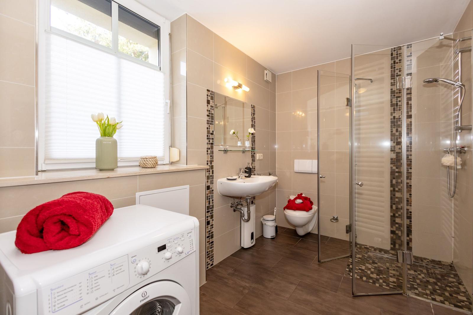 Badezimmer mit Dusche, Waschbecken und Waschmaschine. Fenster mit Blumen.