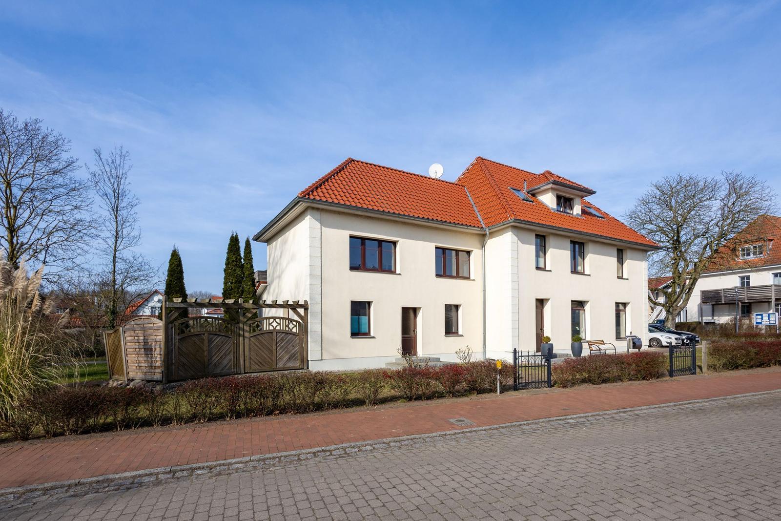 Ein zweistöckiges Haus mit rotem Dach und weißen Wänden. Vor dem Haus befindet sich ein Garten mit Zaun und Bäumen.
