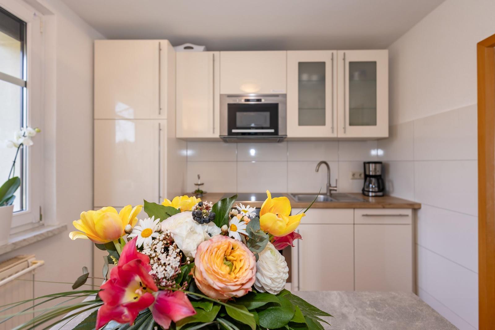 Küche mit weißen Schränken, Spüle und Mikrowelle. Blumenarrangement im Vordergrund.