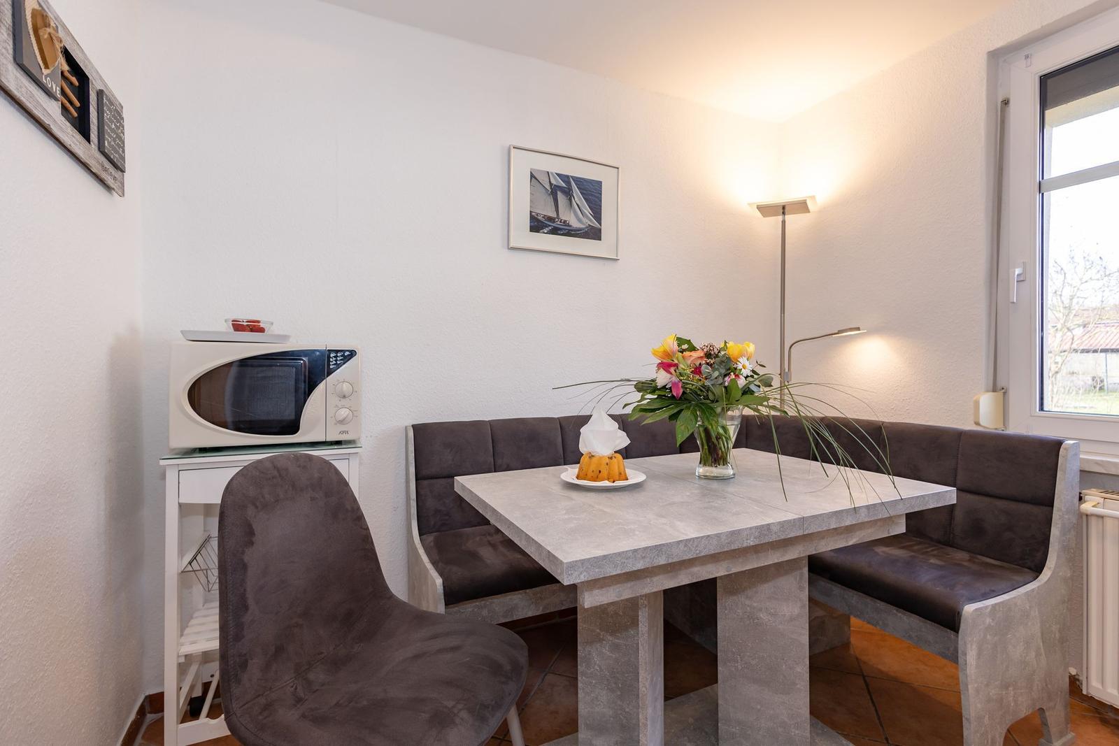 Essbereich mit Tisch, Sitzbank und Stuhl. Mikrowelle und Blumen auf Tisch.