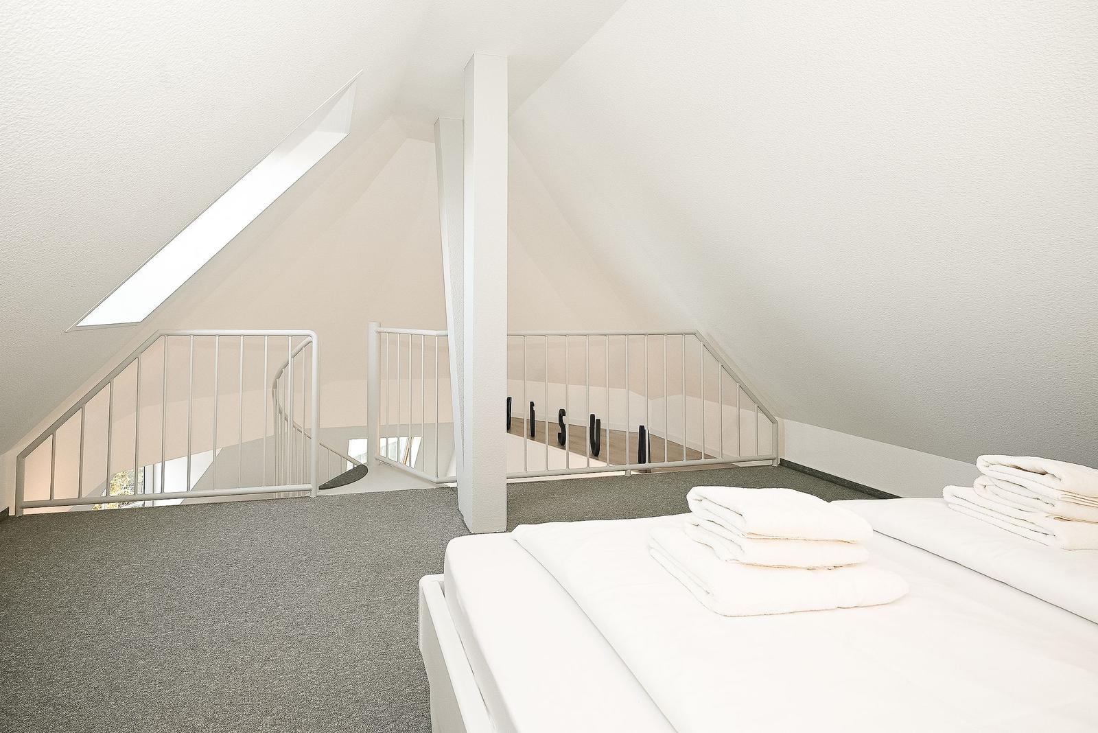 Schlafzimmer mit Dachschrägen, weißem Bett und Dachfenster. Geländer führt zur Treppe.