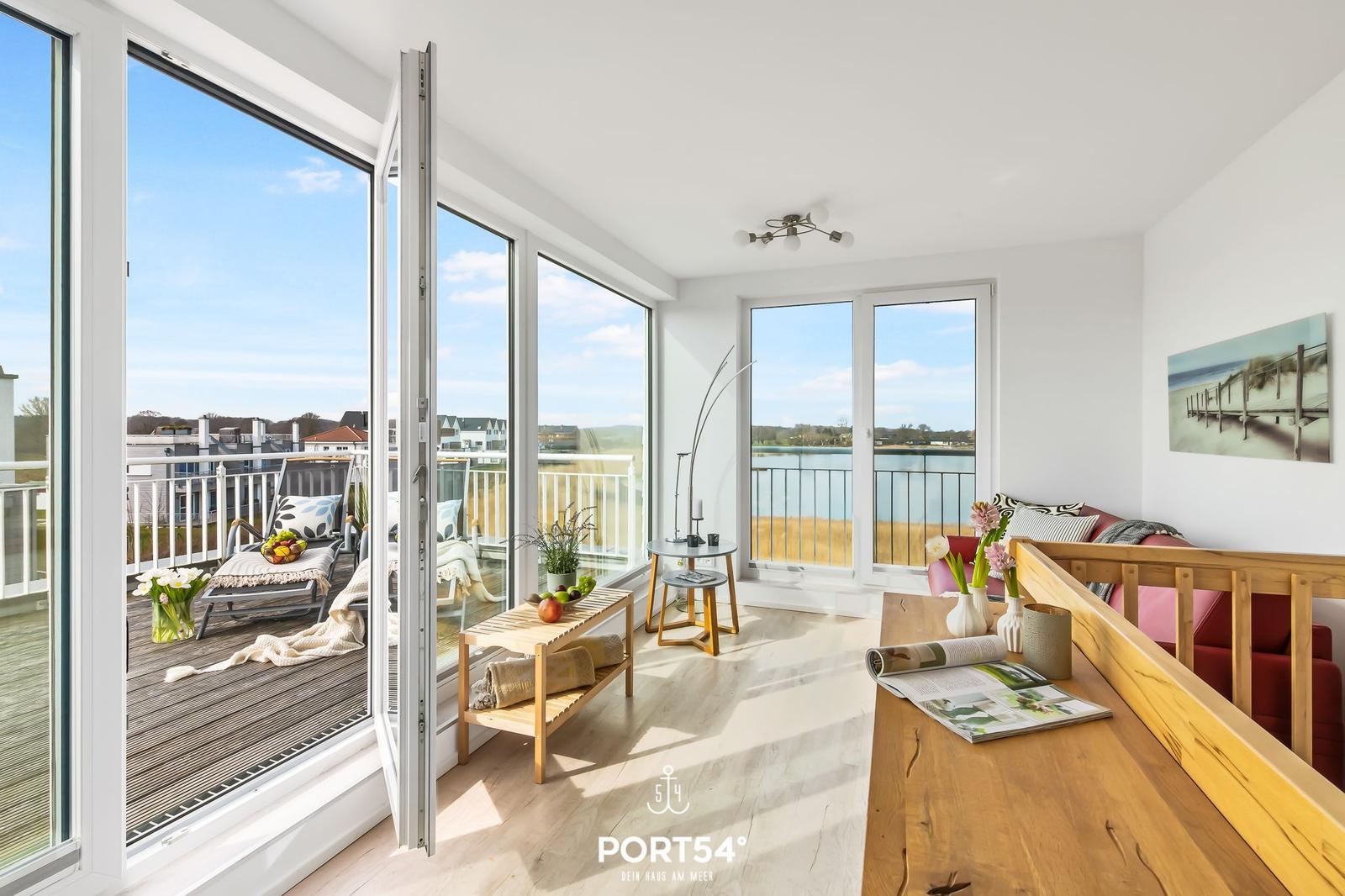 Wohnbereich mit Blick auf Balkon und Wasser. Holztisch, Sofa und große Fenster.