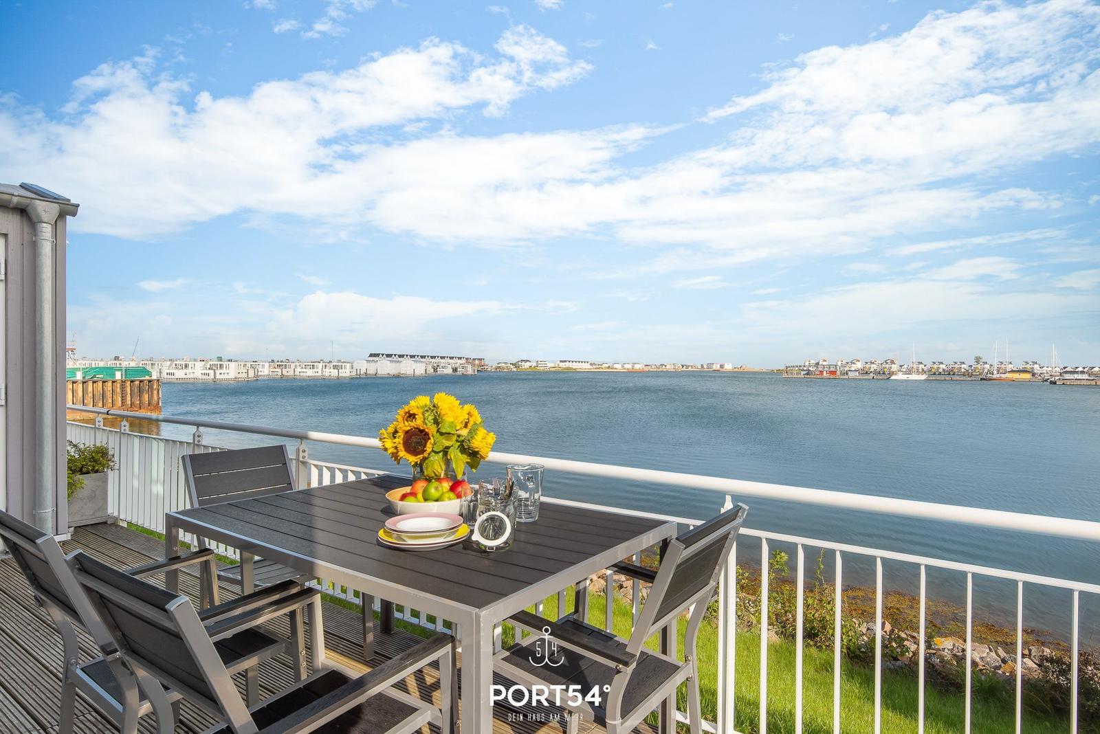 Terrasse mit Tisch und Stühlen, Blick auf Wasser und Hafen.