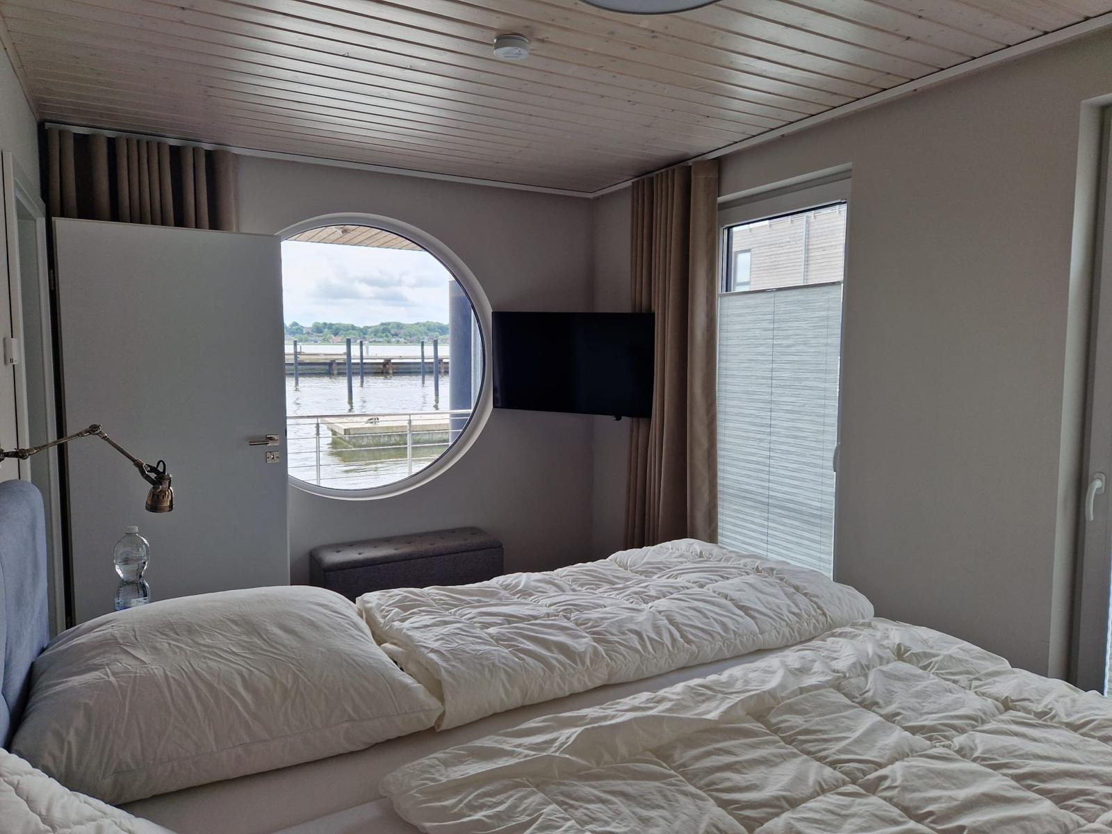 Schlafzimmer mit Rundfenster zum Wasser und Fernseher an der Wand.