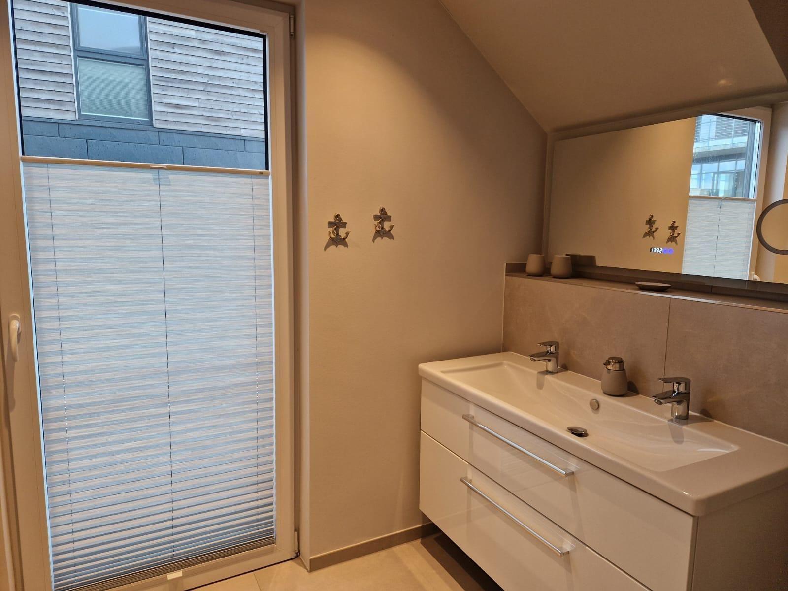 Badezimmer mit Waschbecken, Spiegel und Fenster mit Jalousie.