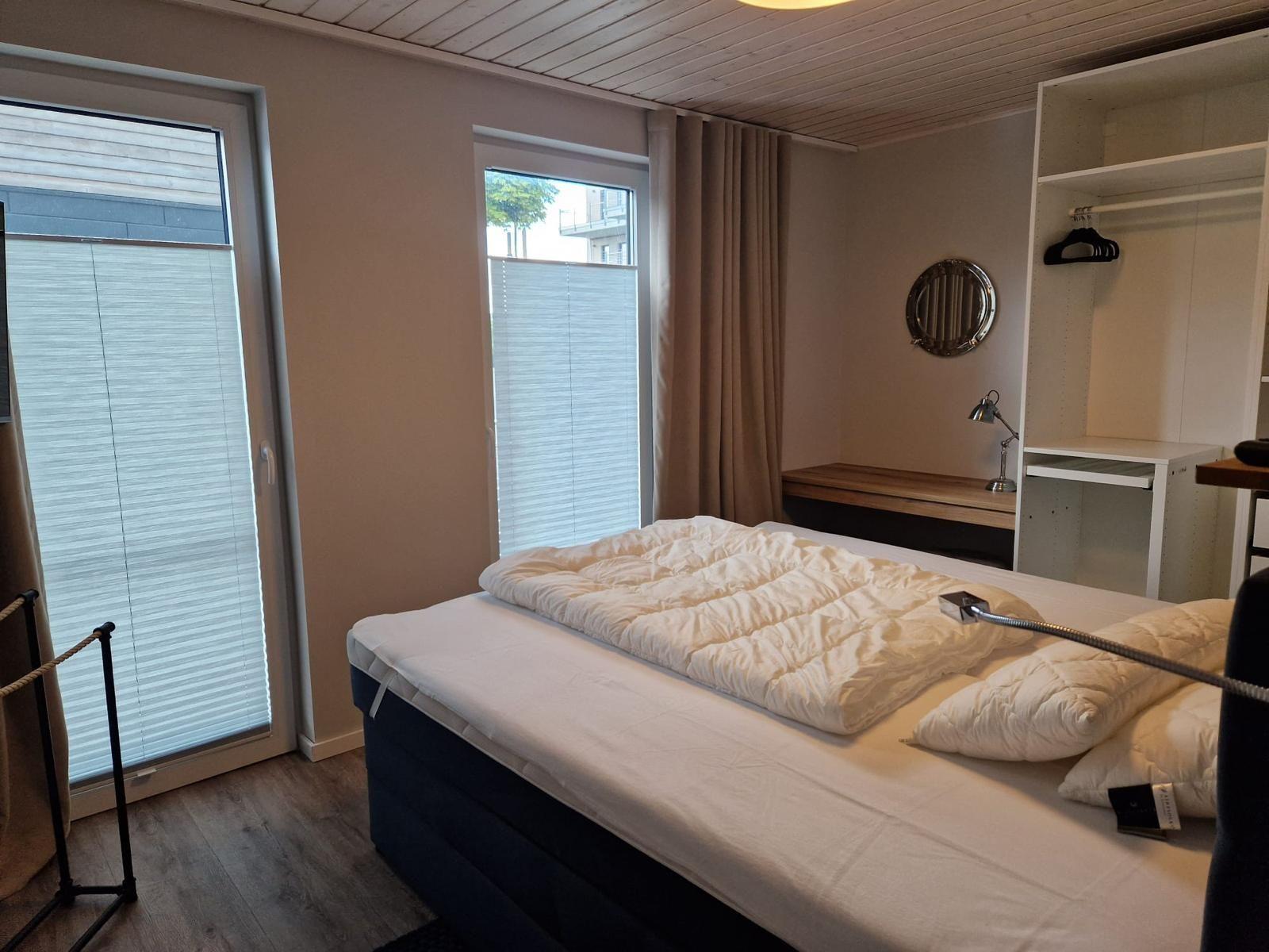 Schlafzimmer mit Doppelbett, Kleiderschrank und Fenstern mit Jalousien.