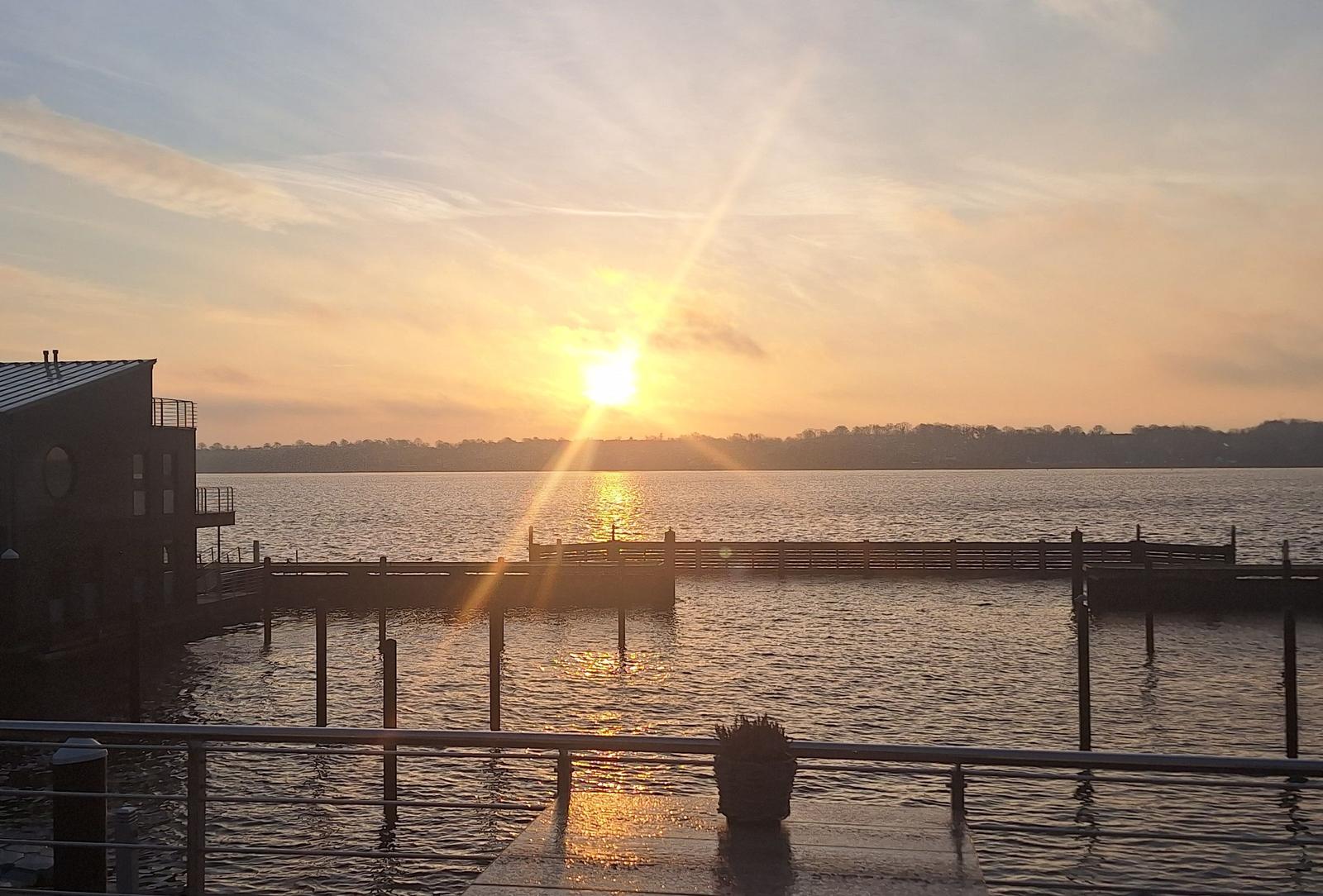 Sonnenuntergang über dem Wasser mit Steg und Gebäude im Vordergrund.