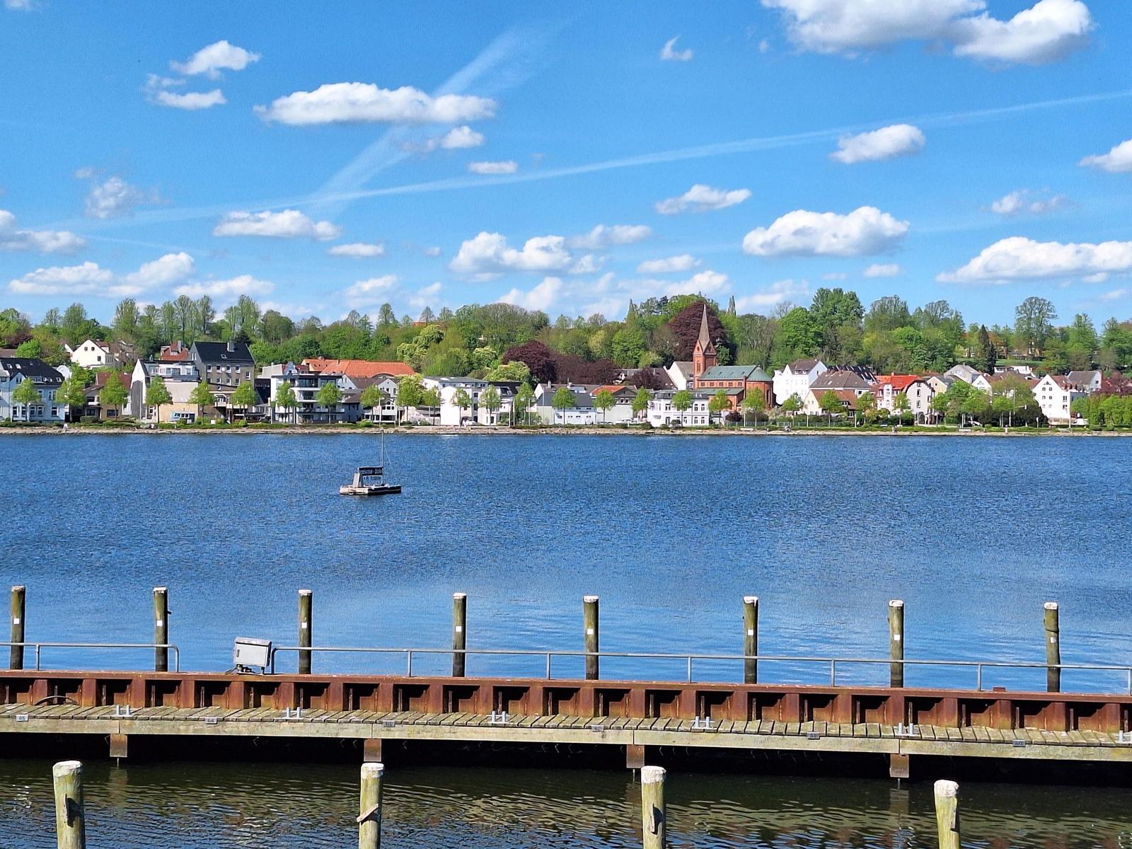 Hafen mit Blick auf einen ruhigen See und eine städtische Uferpromenade mit Häusern und Bäumen.